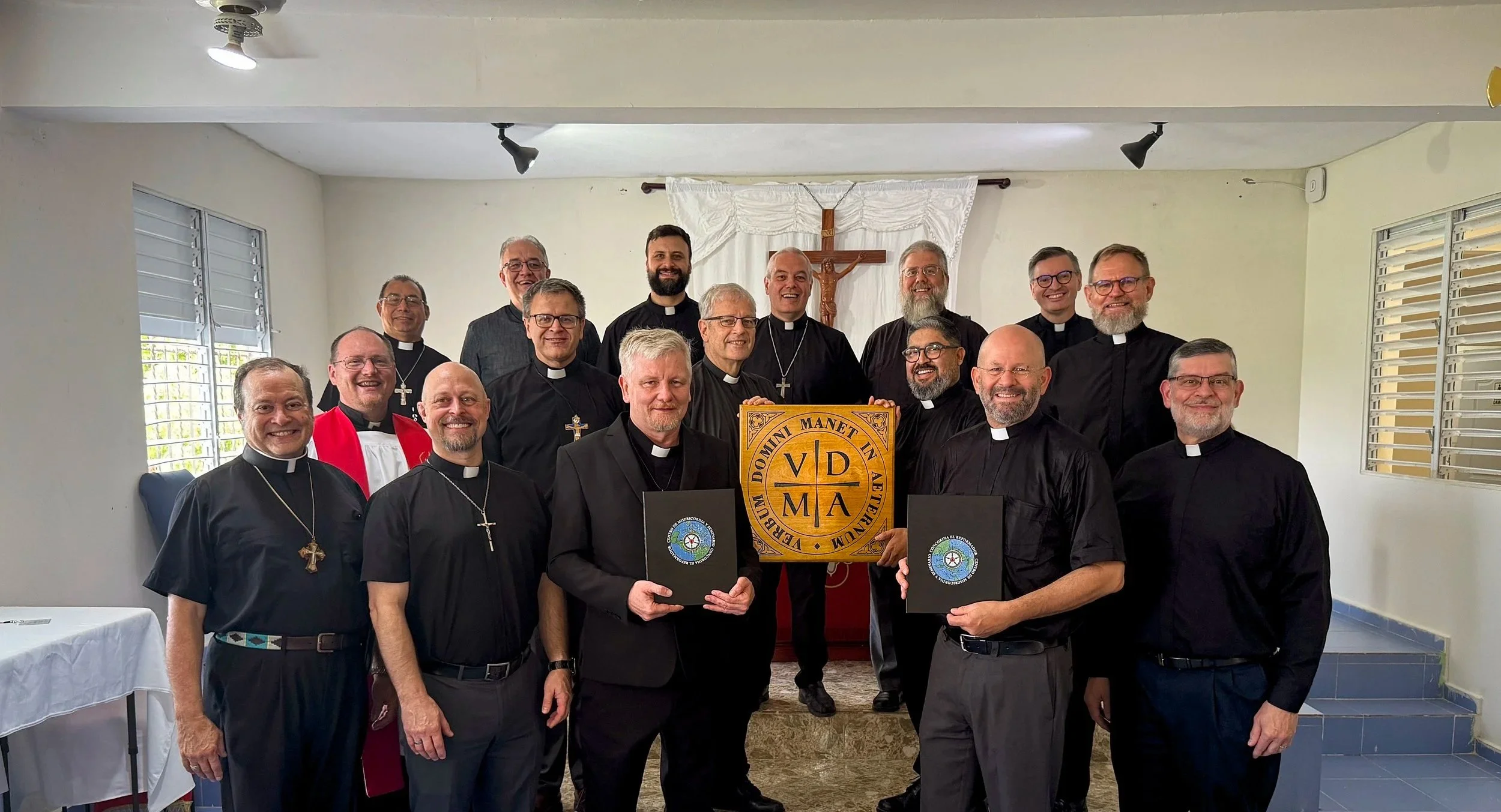 Grupo de religiosos en una ceremonia, algunos sosteniendo documentos y una placa con letras religiosas, en un room con crucifijo al fondo.