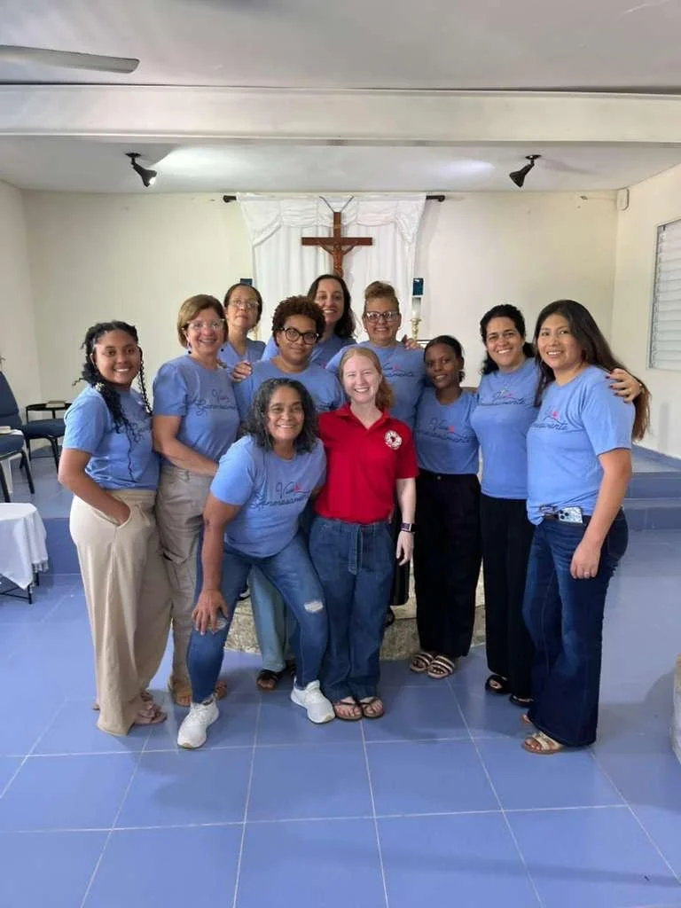 Grupo de personas en una iglesia con cruz en el fondo, varias llevan camisetas azules y una con camiseta roja, todos sonriendo.