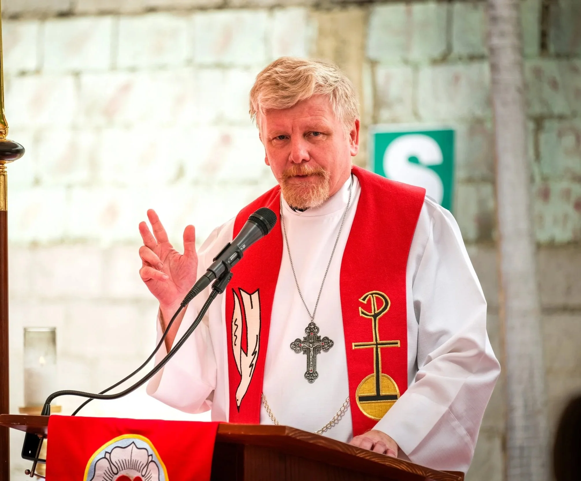 Un pastor luterano en una vestimenta de color blanca y roja, con una cruz colgada en el cuello, hablando frente a un micrófono en un espacio con paredes de ladrillo y una señal verde de salida en el fondo.