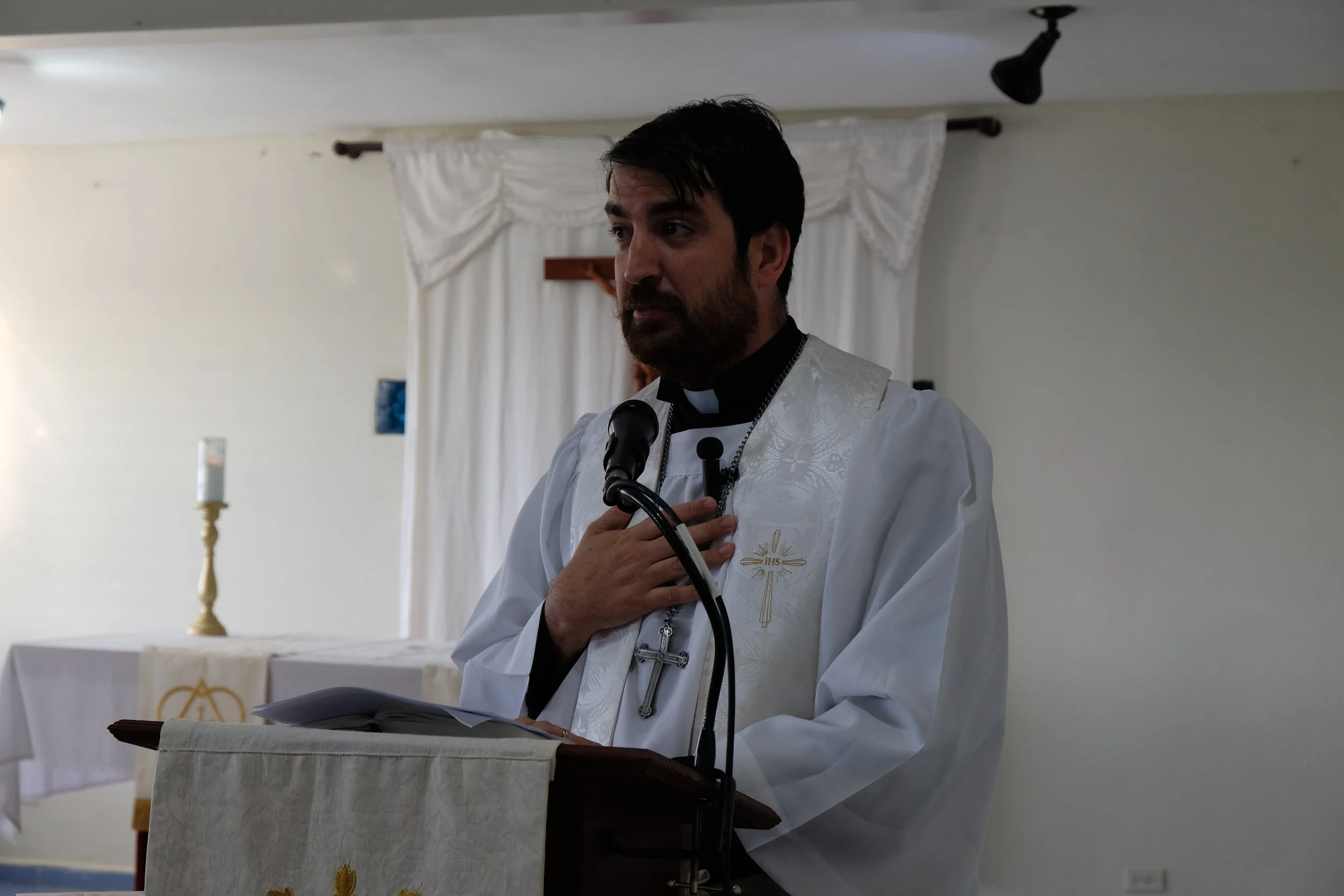 Un pastor en una iglesia luterana sosteniendo la mano en el corazón, vestido con sotana negra y sobre la camiseta blanca con símbolos religiosos, frente a un altar con vela y cruz, en un fondo con cortinas blancas.