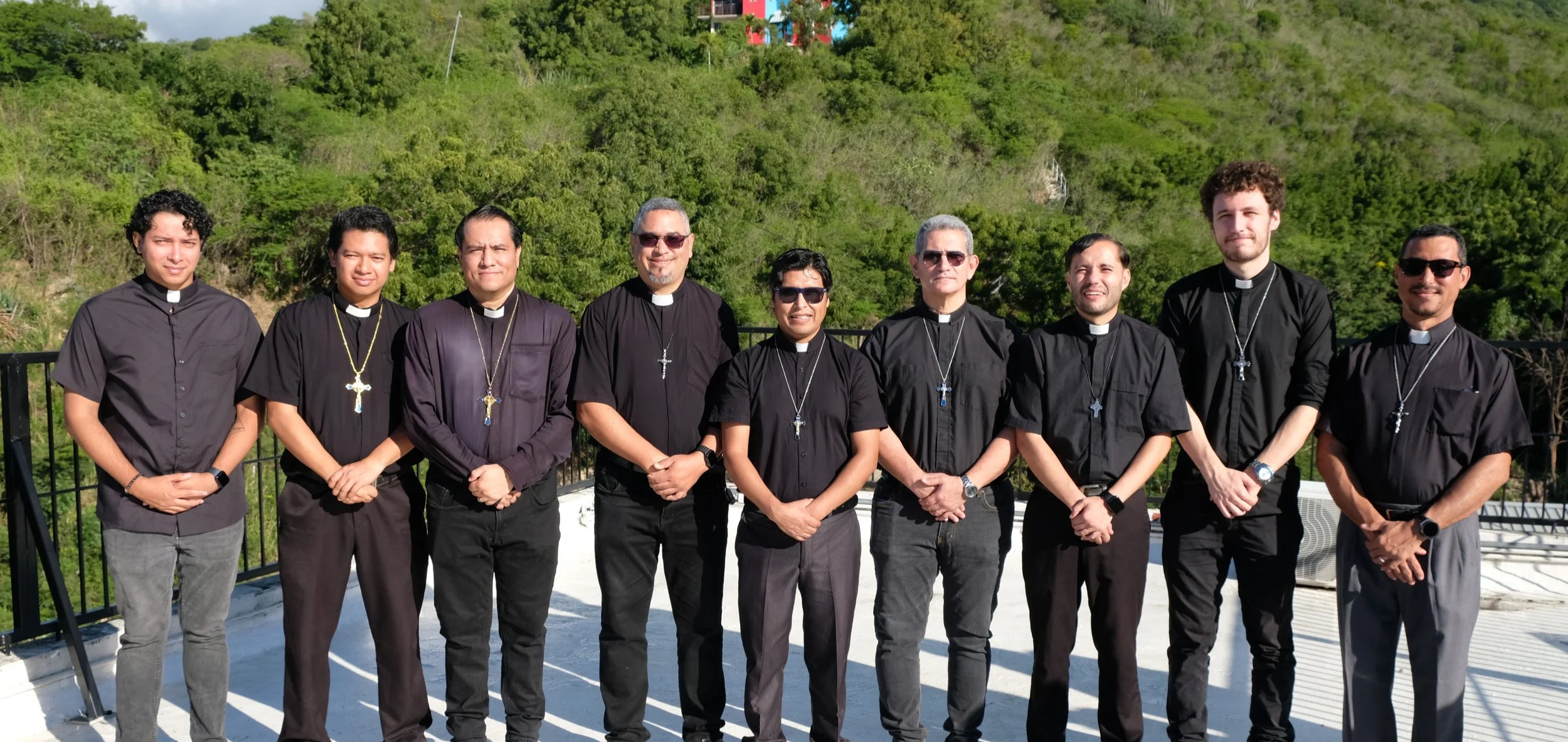 Un grupo de diez sacerdotes con vestimentas negras y cruces colgadas en cadenas, posando al aire libre con fondo de vegetación verde.