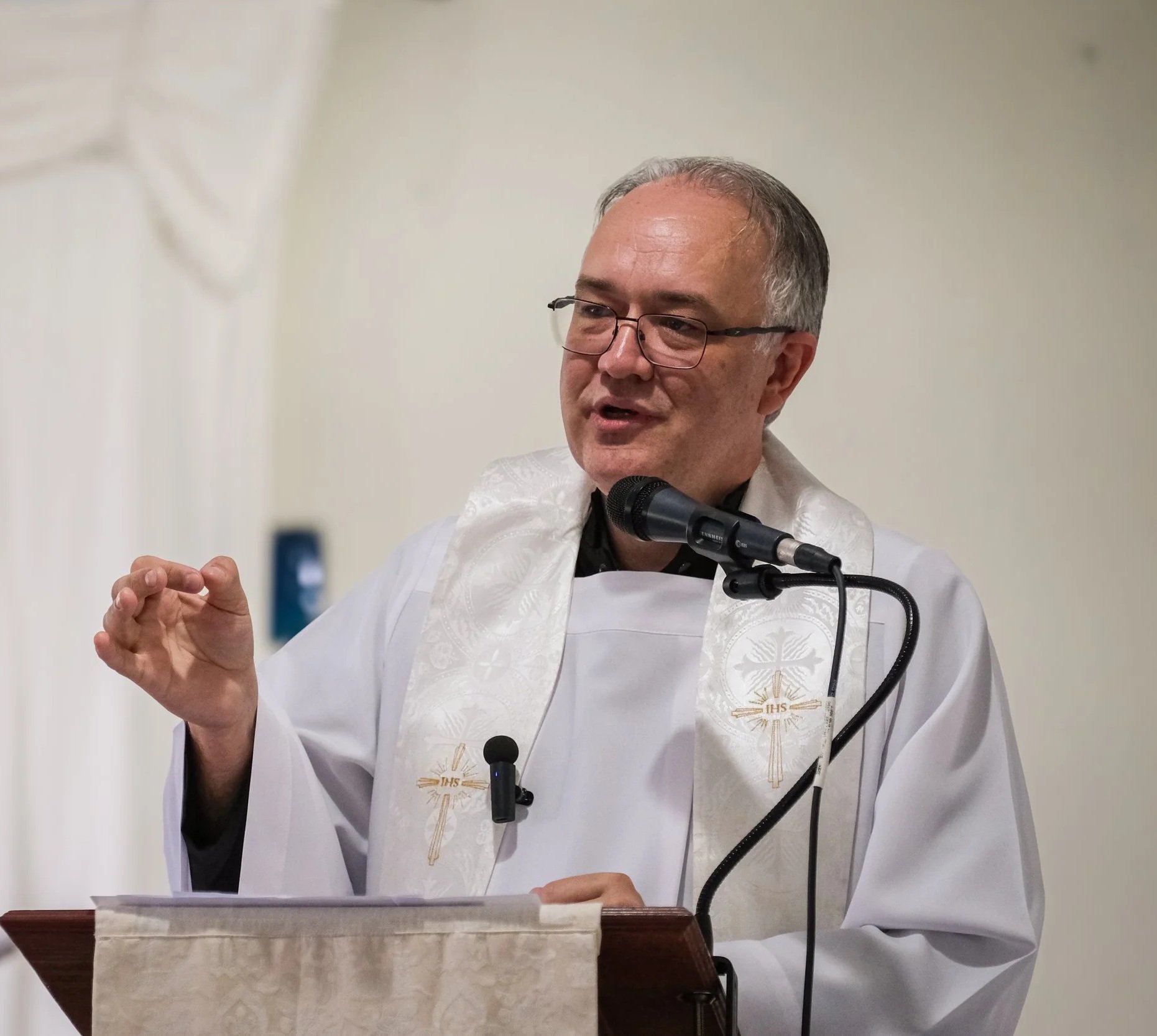 Pastor luterano con gafas hablando en un micrófono, viste ornamento litúrgico blanco con símbolo religioso.