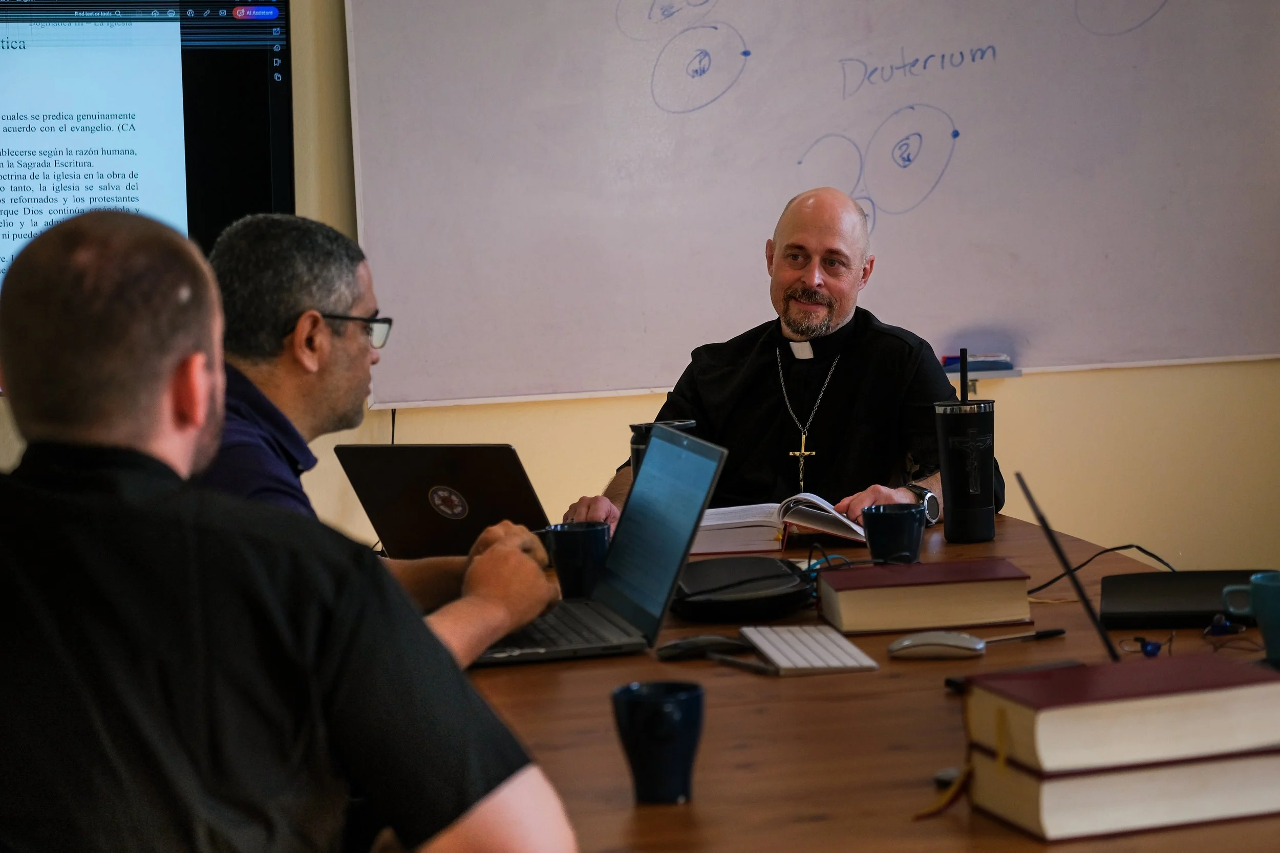 Un grupo de personas, incluyendo un sacerdote con collar y cruz, participan en una reunión en un aula, con una pizarra y una pantalla con texto visible en el fondo.