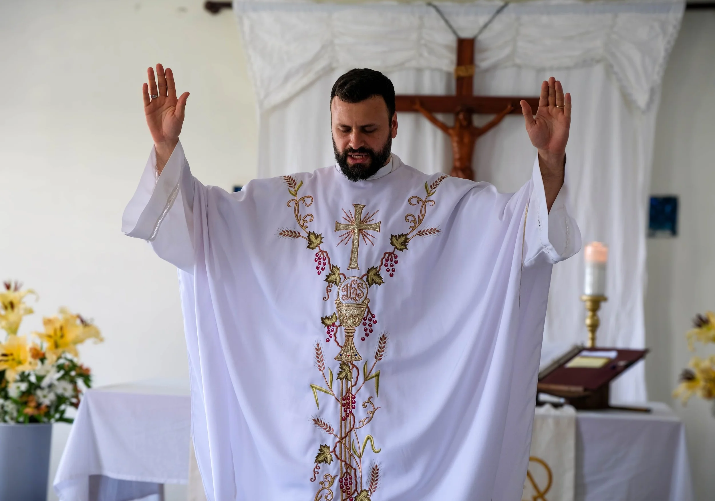 Sacerdote católico en misa con brazos levantados, vestido con alba decorada con símbolo de la cruz y motivos religiosos, en interior de iglesia.