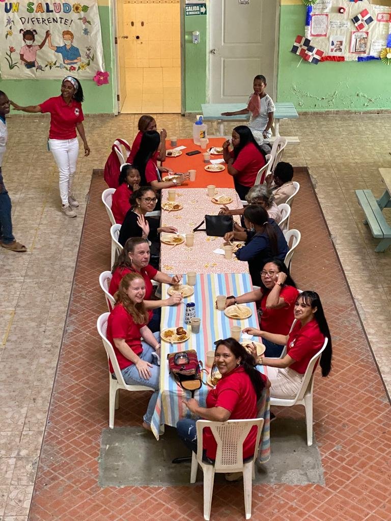 Un grupo de mujeres y niños sentados en una mesa celebrando, algunas sonriendo y disfrutando de comida, en un medio ambiente escolar con decoraciones y un cartel que dice 'Un saludo diferente' en un salón con paredes verdes.