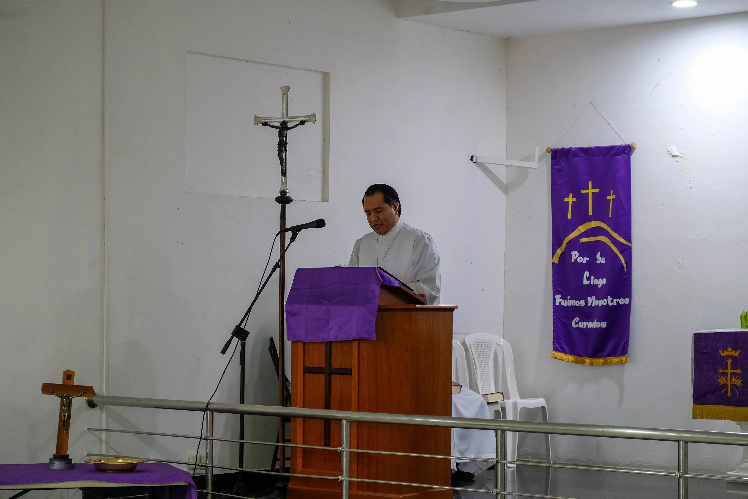 Un sacerdote está de espaldas, frente a un atril en una iglesia, con un crucifijo en la pared y un banner morado con cruces y texto. Hay sillas blancas y una mesa cubierta con tela morada.