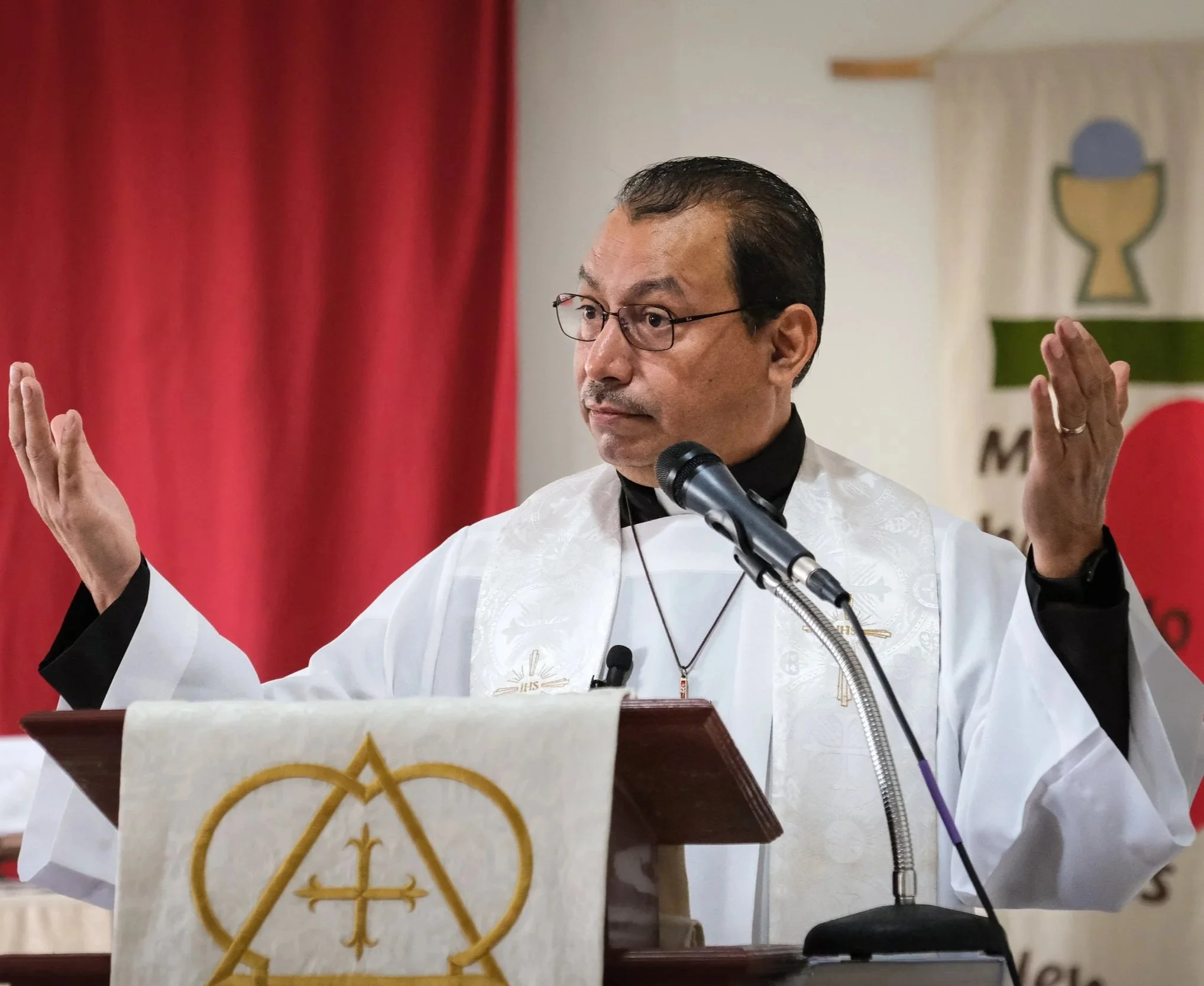 Un pastor con gafas y barba, vestido con sotana y capa blanca, está en un podio durante un servicio luterano, con un fondo rojo y un símbolo de una copa en la pared.
