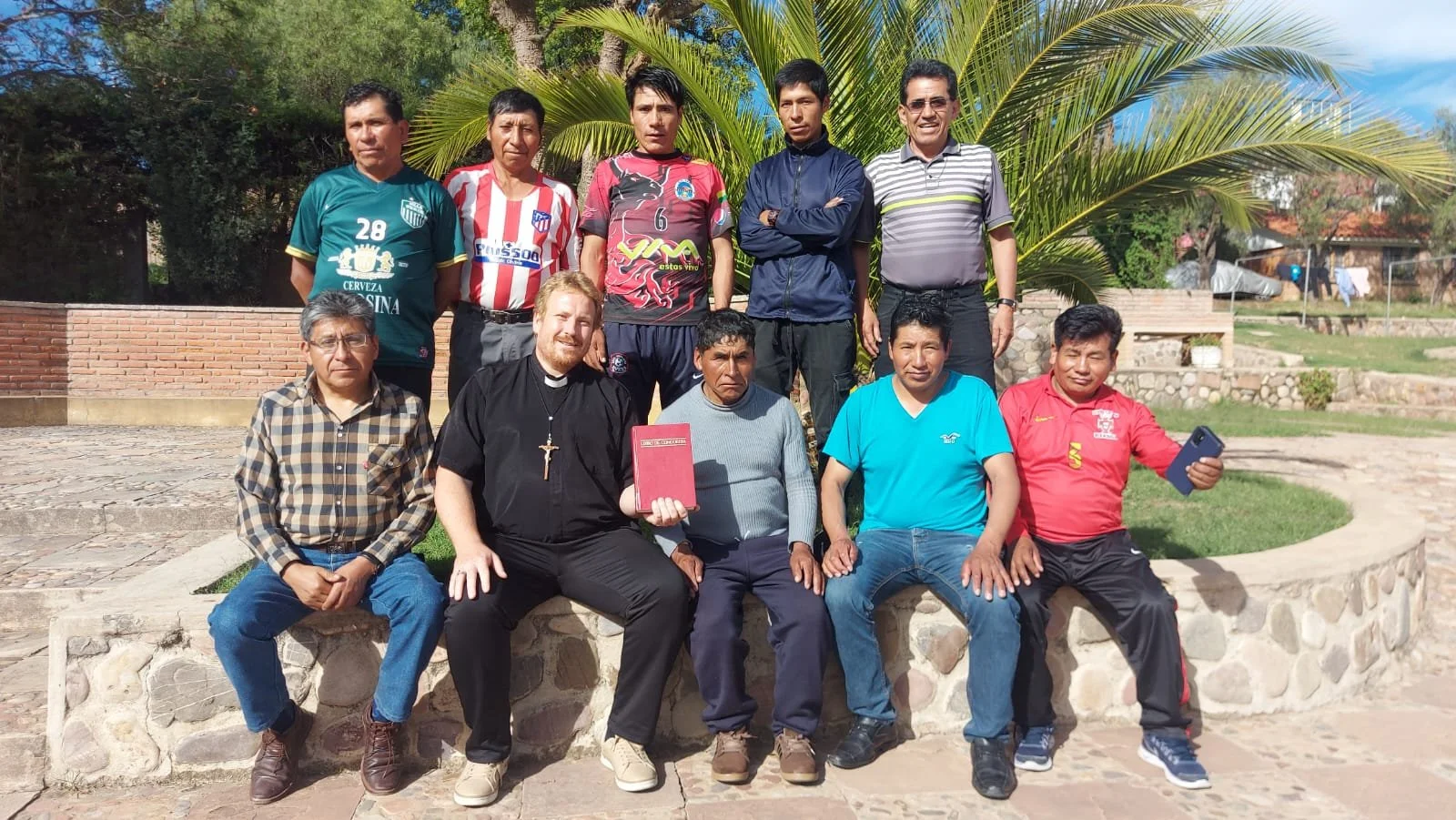 Grupo de once personas posando en un parque con plantas y áreas verdes, algunos con vestimenta casual, algunos con camisetas de fútbol, una persona con vestimenta clerical, en un día soleado.