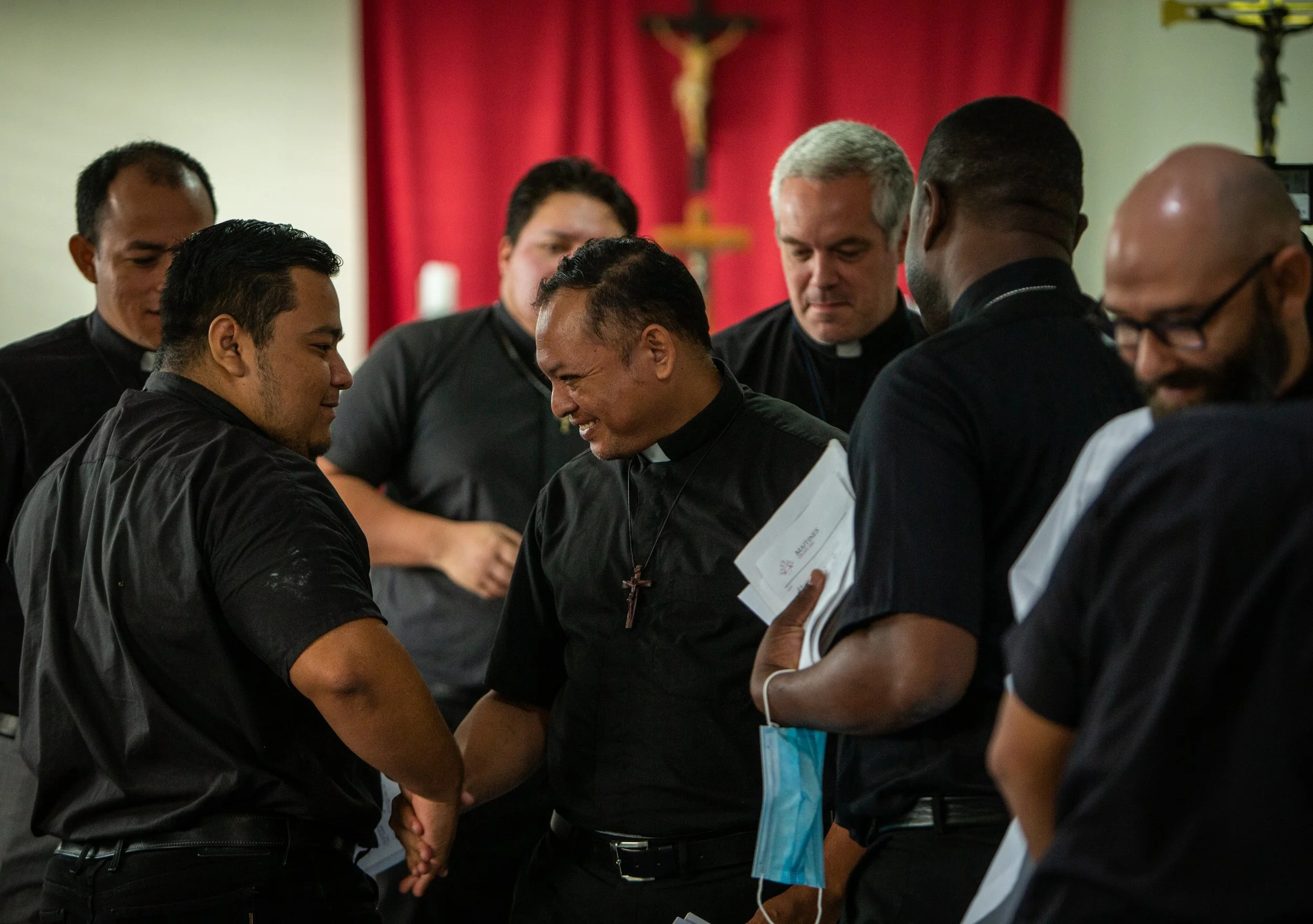 Grupo de personas, en su mayoría pastores luteranos, dándose la mano en una ceremonia religiosa, en un lugar con cortinas rojas y una cruz en el fondo.