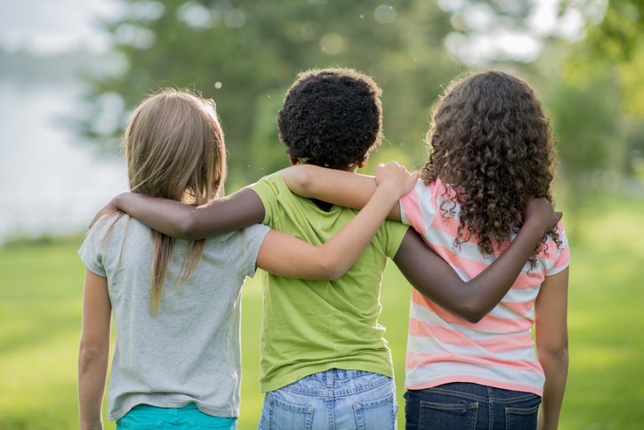 Three children standing outdoors with their arms around each other, facing away from the camera, in a grassy park with trees.