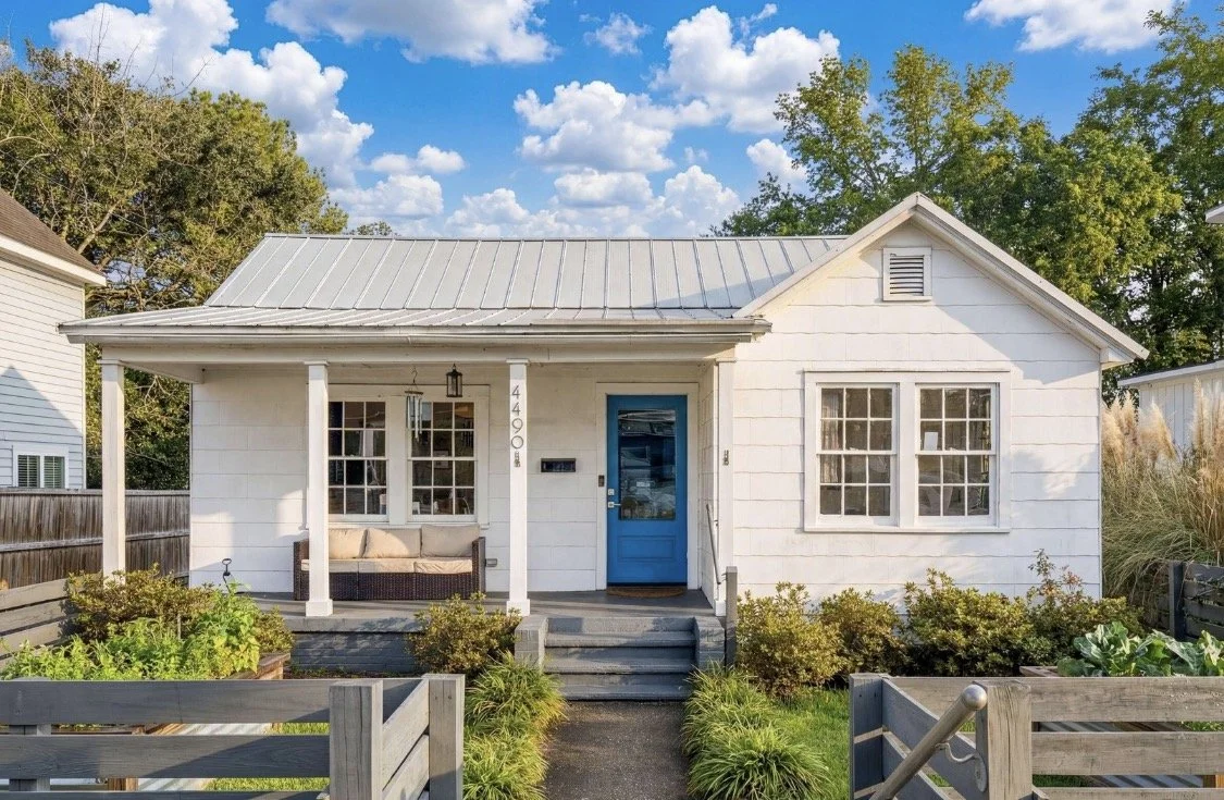 Front view of a small white house with a metal roof, blue front door, and a porch with a bench. There are garden beds and greenery in front, with trees and a partly cloudy sky in the background.