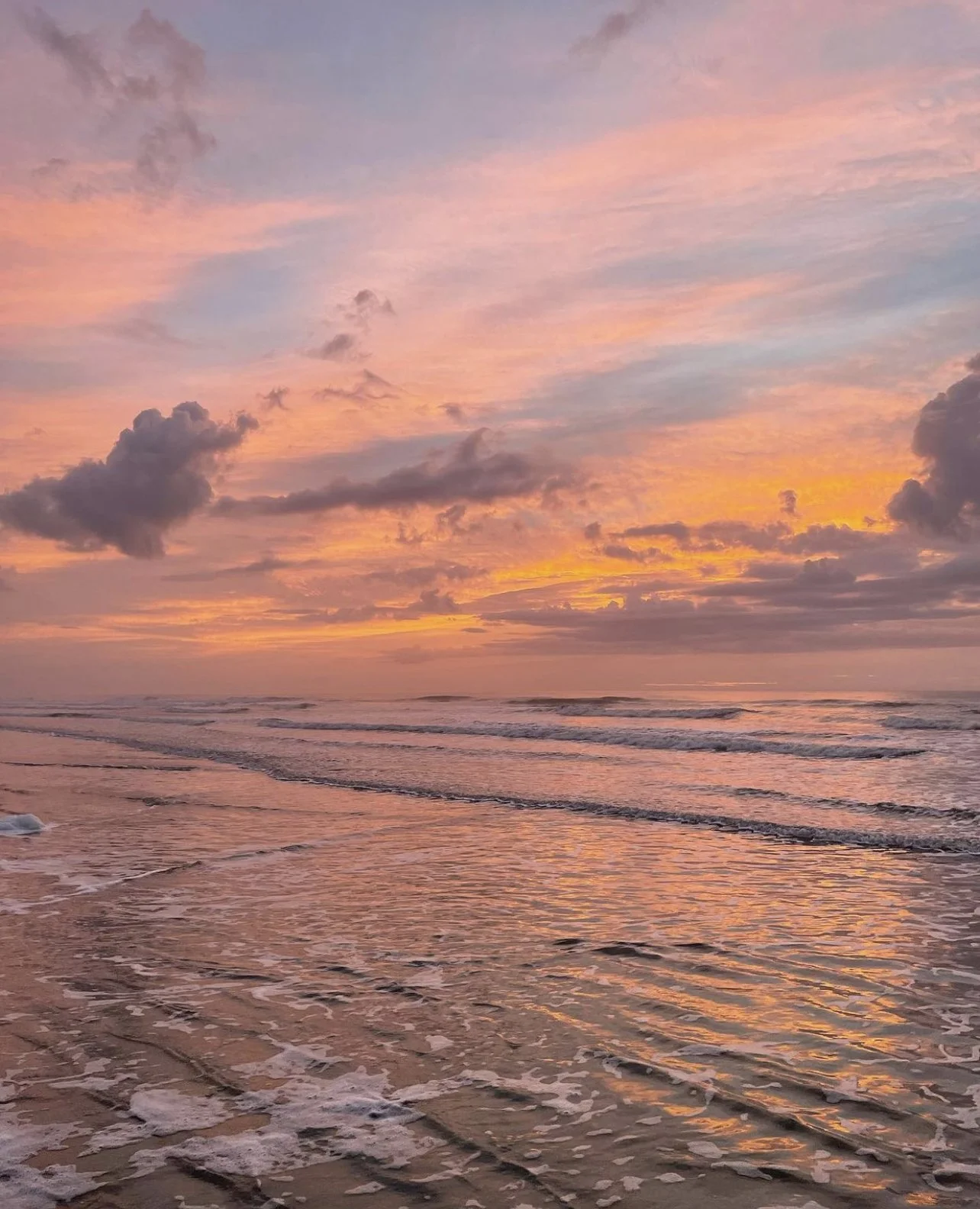 A beach at sunset with pink, orange, and purple clouds in the sky and gentle waves lapping the shore.