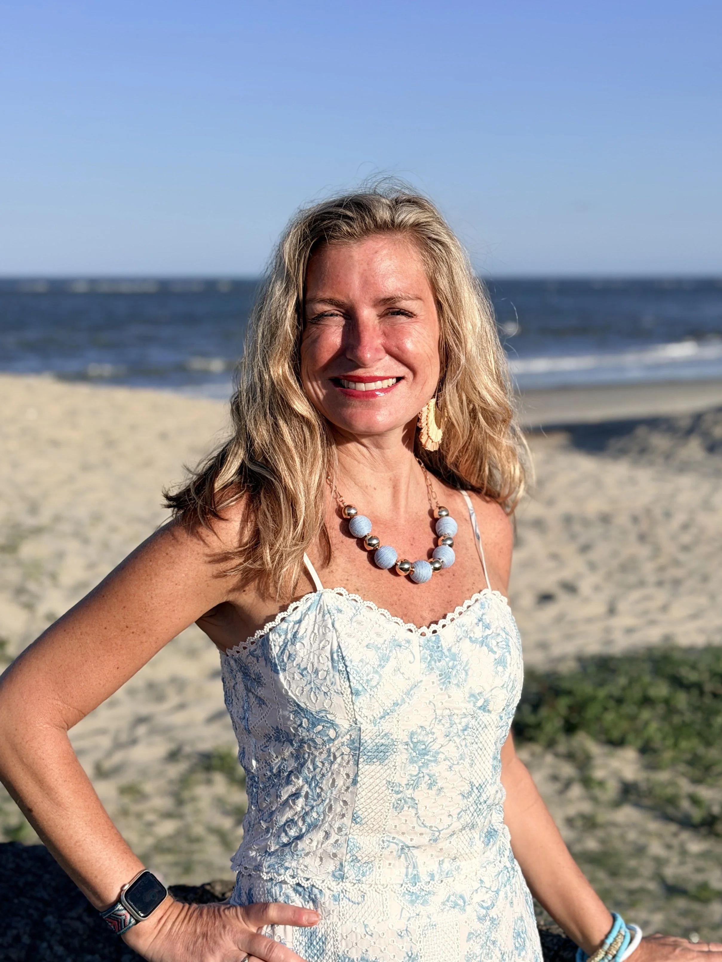 Woman smiling on the beach with ocean and blue sky in the background, wearing a white lace dress and jewelry.