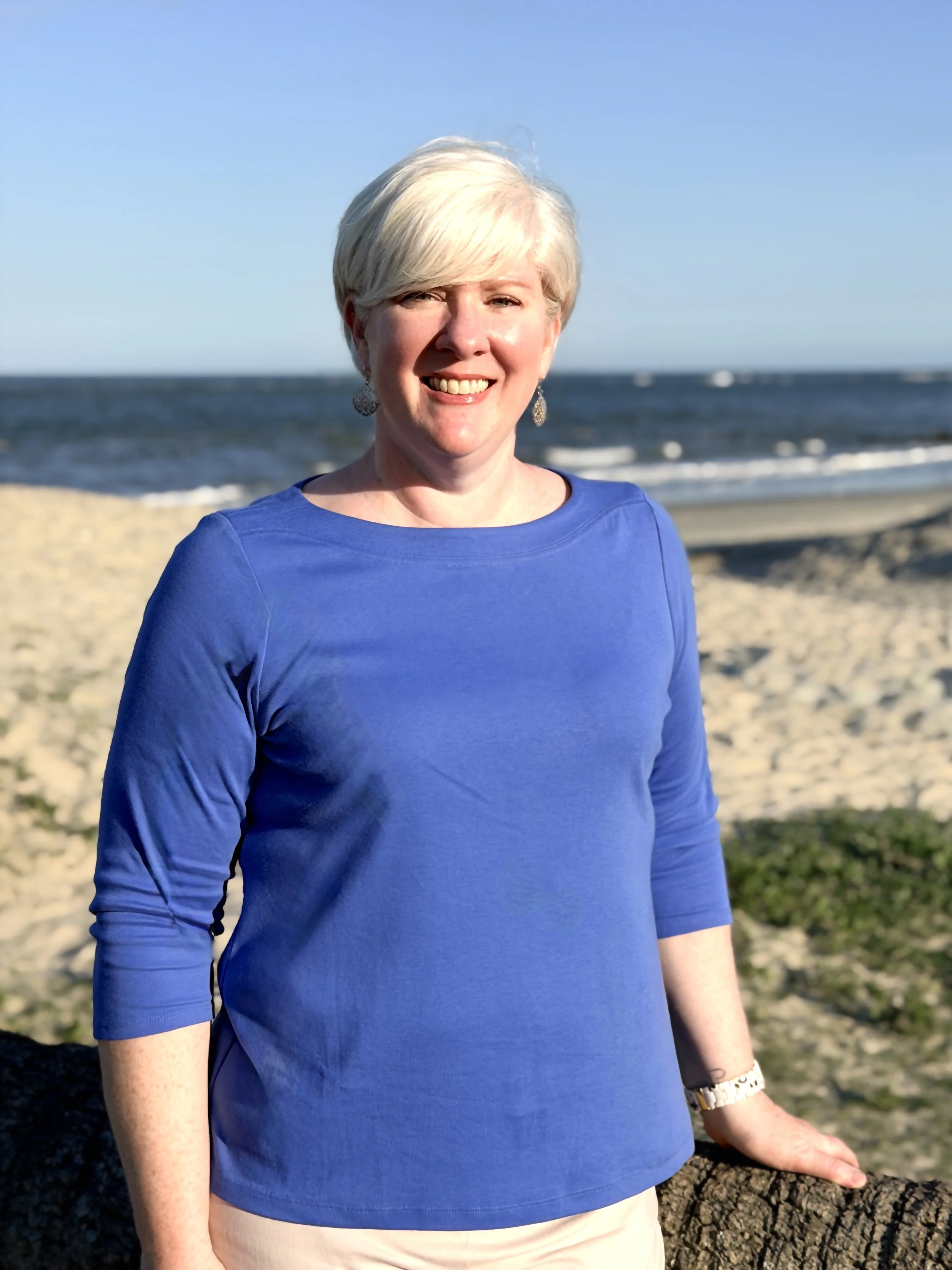 A woman with short white hair smiling on a beach, wearing a blue top, earrings, and a watch, with the ocean and sandy beach in the background.