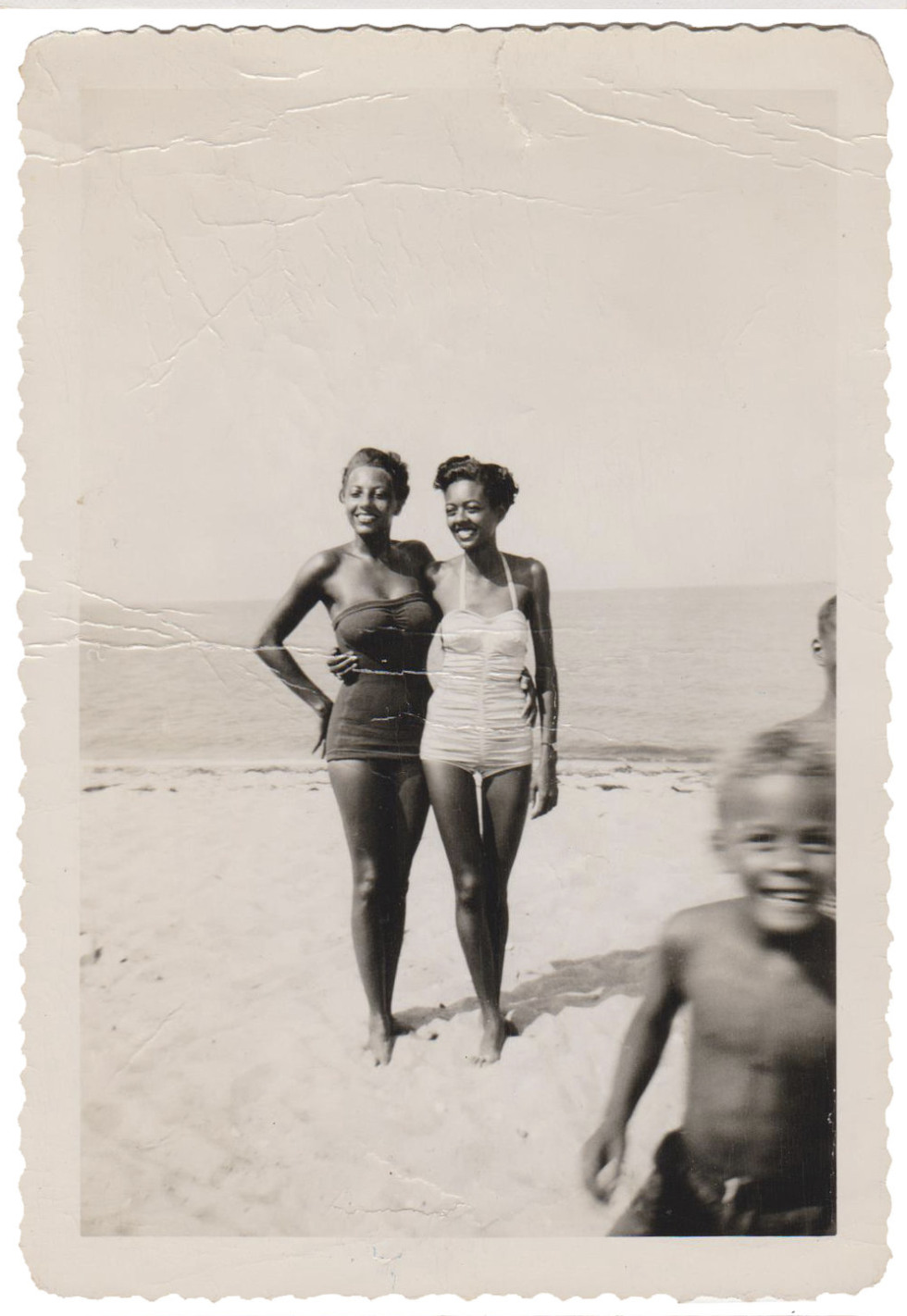 Two women standing on a beach, smiling, in swimsuits, with the ocean in the background, and a young boy in motion in the foreground. Circa 1955.