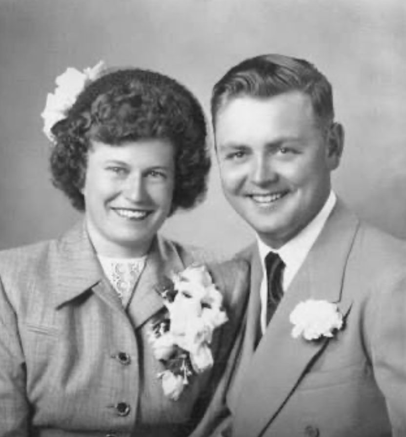 Black and white engagement portrait of a smiling woman and man from the 1940s, dressed in vintage formal attire, with flowers on their clothing.