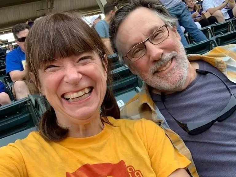 Elizabeth Meister, with brown hair in pigtails and a "No Lights At Wrigley" shirt sitting in Wrigley Field in Chicago, with her husband Dan Collison, bearded with glasses.