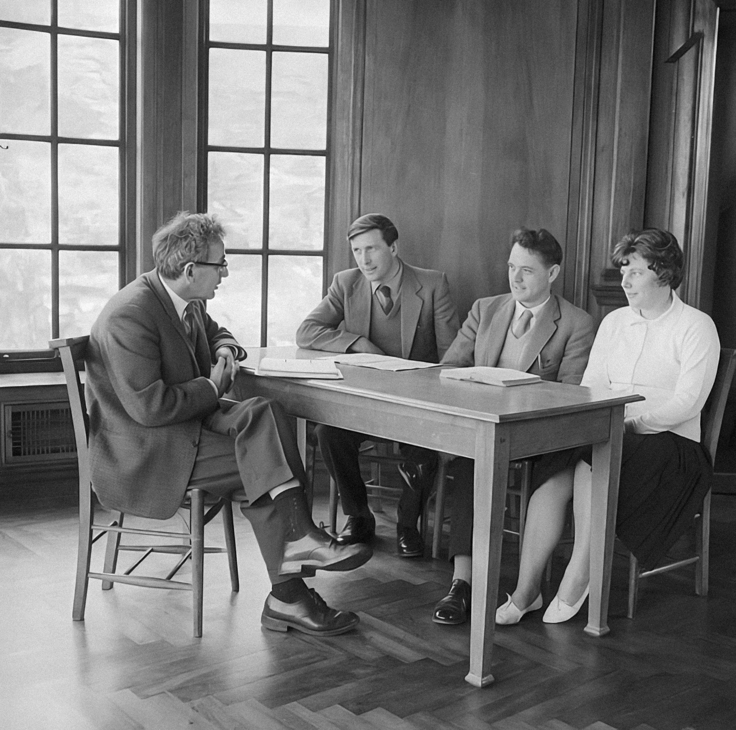 Black and white photo of four men sitting around a table in a room with large window viewing the sky, engaged in conversation.