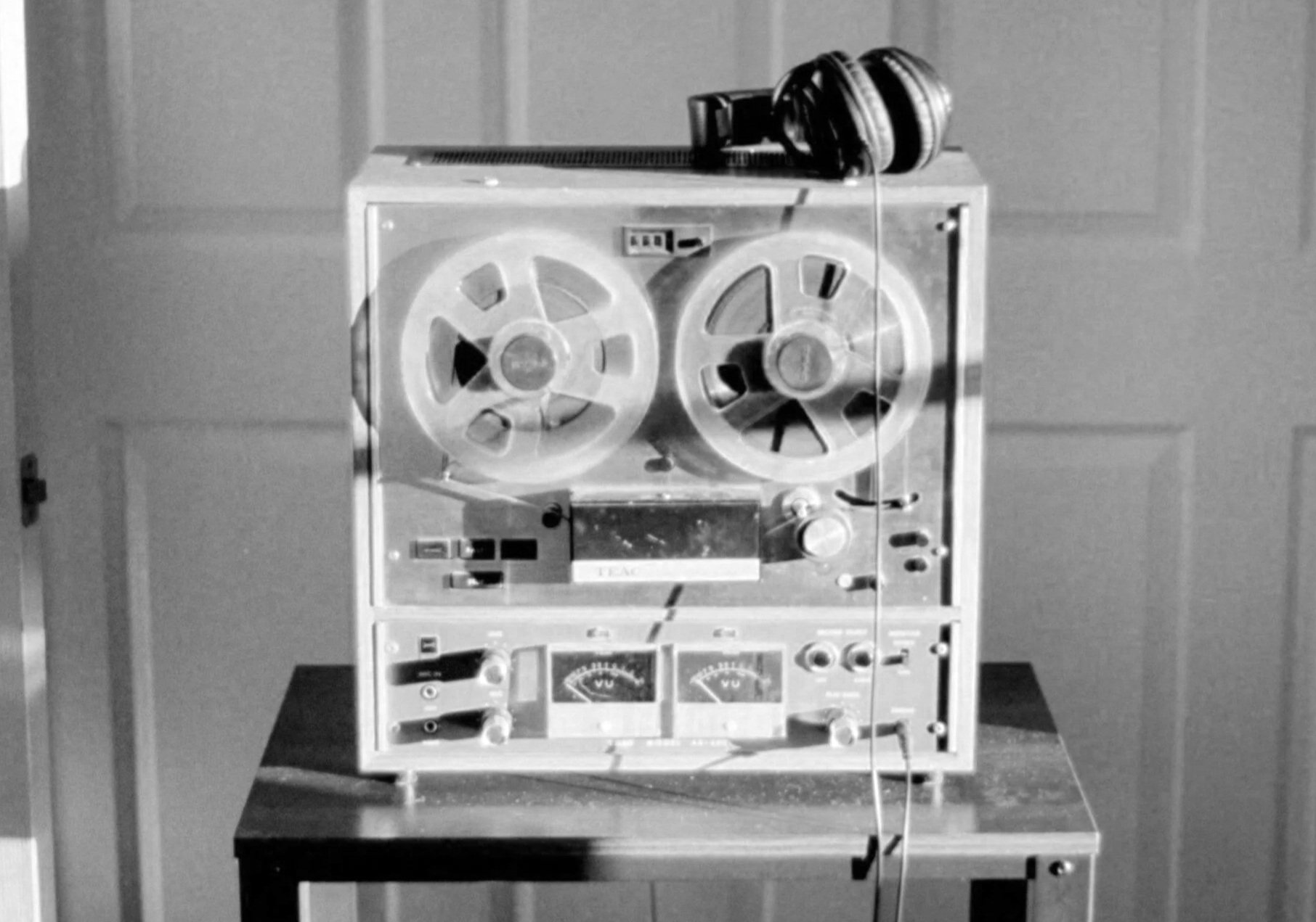 Black and white photograph of a vintage reel-to-reel tape recorder on a table. The tape recorder has two large reels with tape wound around them, various knobs, and meters. A pair of over-ear headphones rests on top of the recorder.