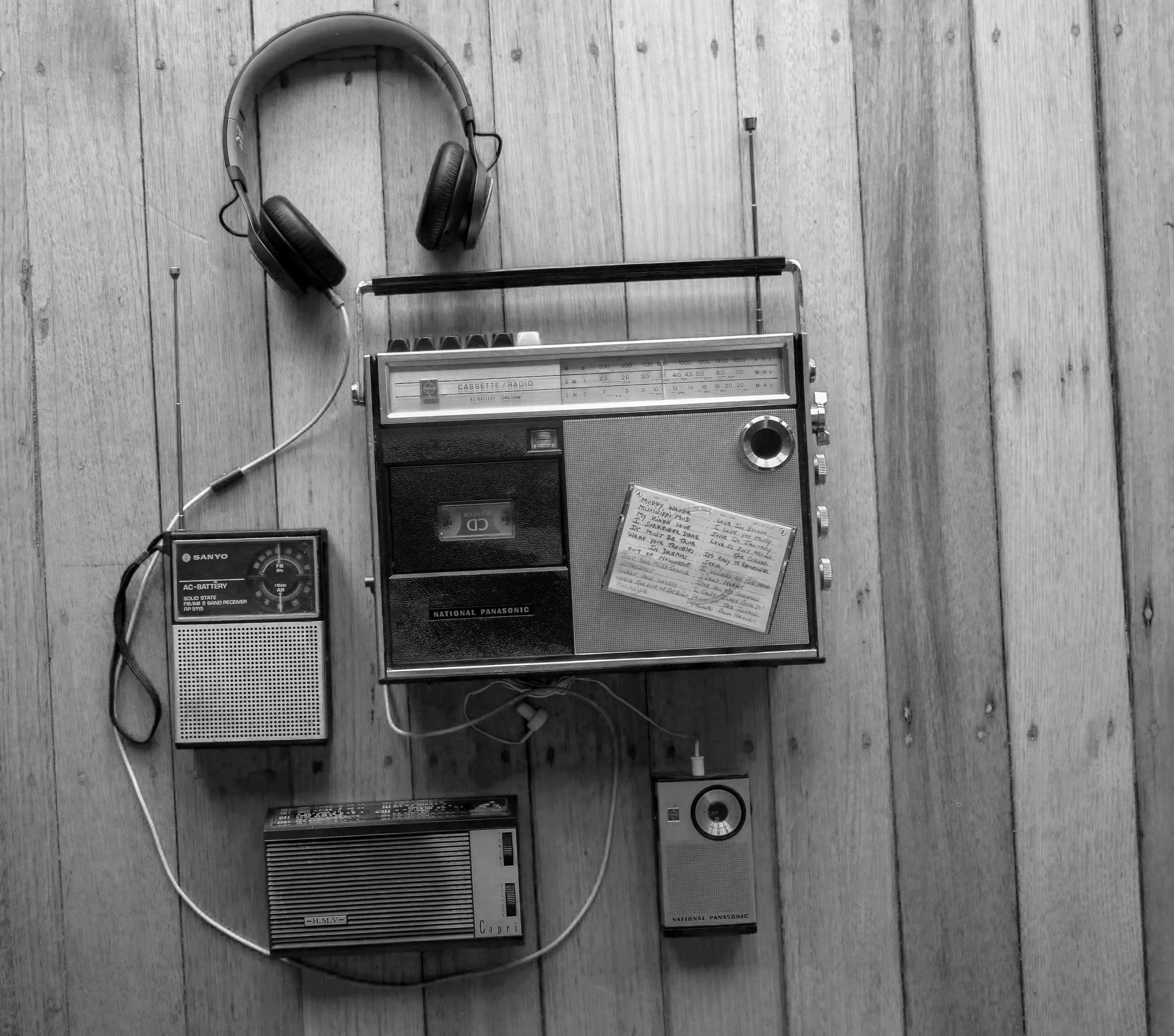 Black and white photograph of vintage radios and headphones placed on a wooden floor.