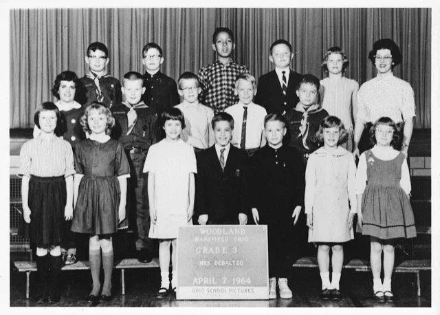 A black-and-white photo of the Third Grade class from Woodland Elementary School in 1964, featuring children and a teacher, arranged in three rows on stage with curtains in the background. Dan Collison is front row center.