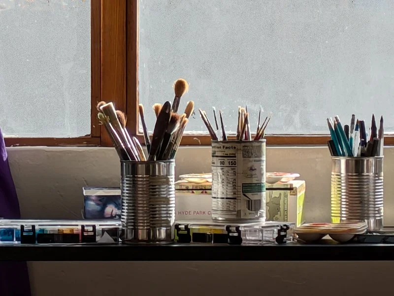 A window sill with three tin can containers holding paintbrushes, markers, and colored pencils, with boxes and art supplies on a black surface in front.