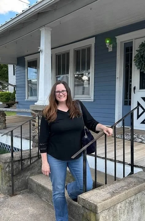 Realtor Lisa Nanni with glasses and long brown hair, wearing a black V-neck and jeans, standing outside a blue house, holding a black purse, on a small concrete porch with a black metal railing.