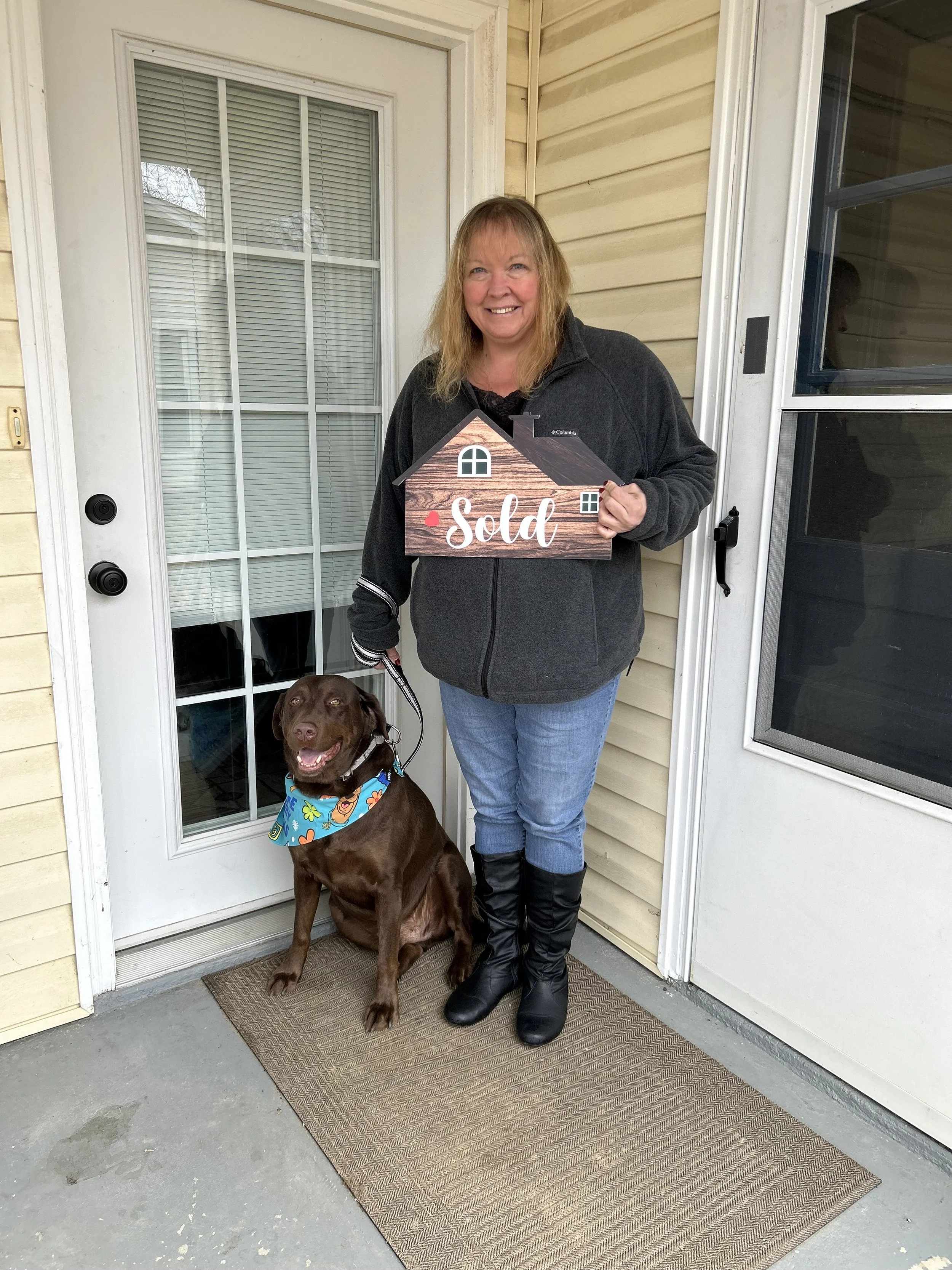 Happy client Sherry, holding a house-shaped sign that says 'sold' stands next to Hershey, her chocolate Labrador dog wearing a colorful bandana, on a porch in front of a house with beige siding and a glass door. Hershey also looks like he's smiling!