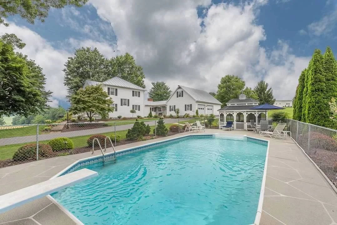 Residential backyard with a swimming pool, white 1910 farmhouse, gazebo, and lush green trees under a brilliant blue sky with soft clouds.