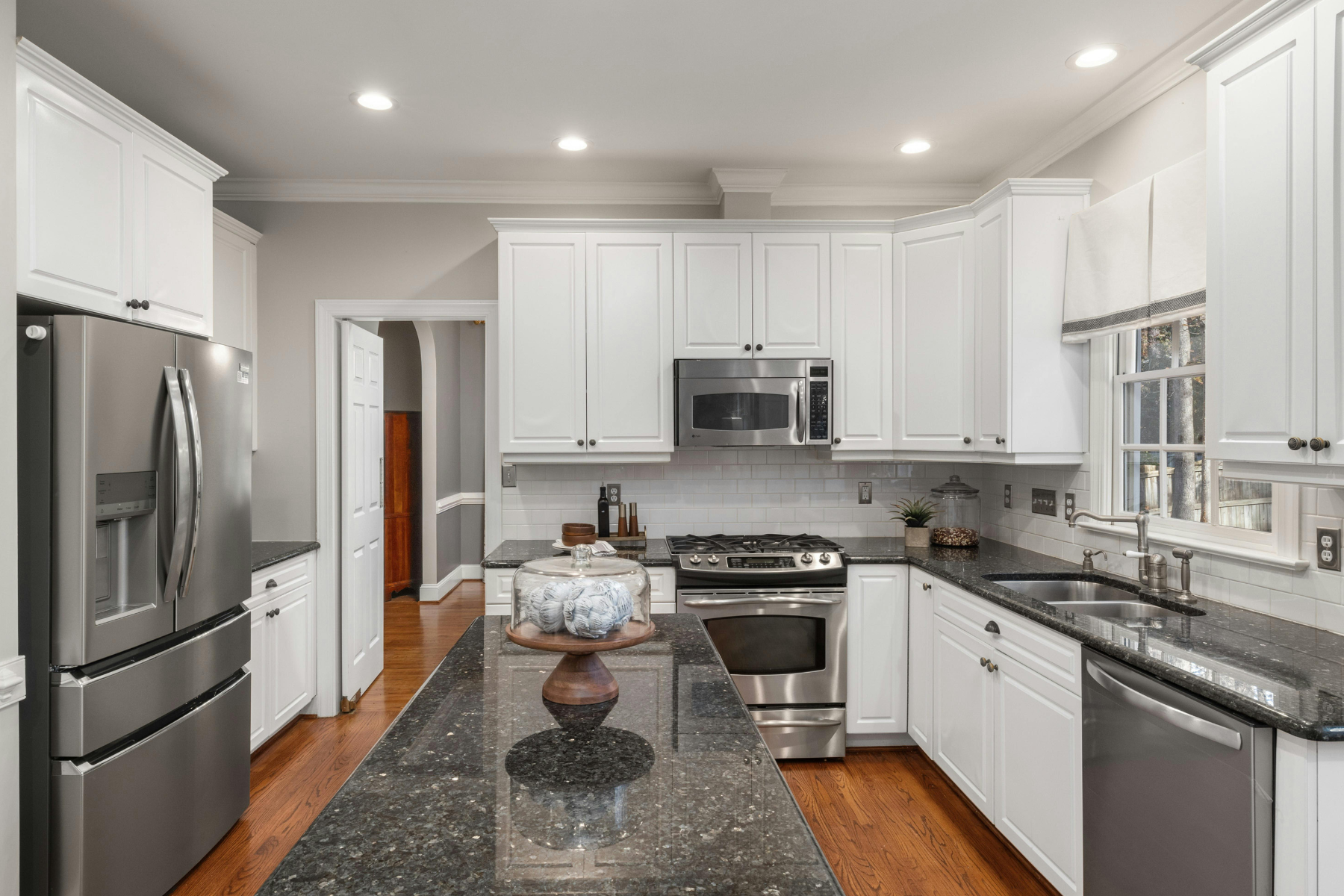 Modern kitchen with white cabinets, black granite countertops, stainless steel appliances, and a center island with a glass cake stand. Includes a window with a white shade and hardwood flooring.