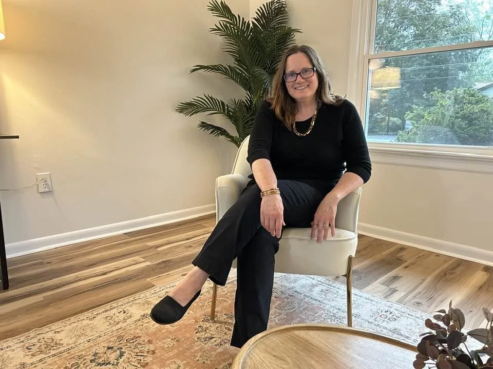 Lisa, with glasses and shoulder-length hair in a staged house - comfortably seated on a white armchair in a well-lit room with wooden floors, a large window, a patterned rug, a plant, and a small table.