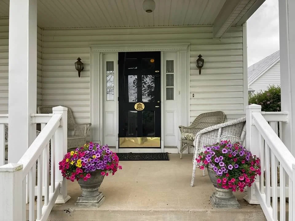 Inviting front porch with white railing, black front door with a gold letter, two wicker chairs, and two large flower pots with pink, purple, and yellow flowers.