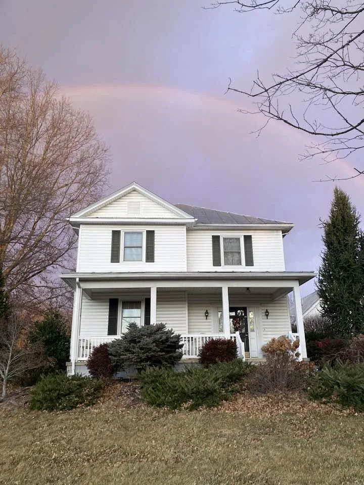 A white two-story four-square house with black shutters and a front porch, surrounded by shrubs and trees, with a visible rainbow in the sky, appearing directly over the house.