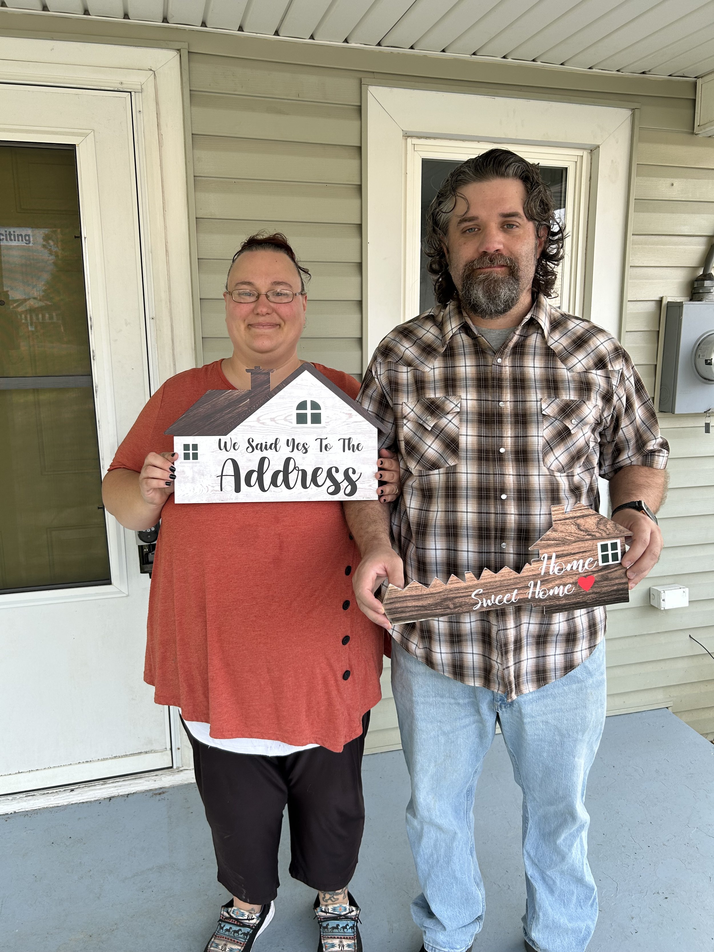Happy clients Matthew and Ashtin, each holding a sign (one is house shaped, the other a key shape) on the front porch of their new home.