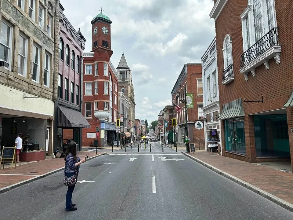 Realtor Lisa is alone, standing profile on the street,  in the middle of downtown historic Beverley Street, with vintage-style buildings lining both sides of the street, a red clock tower in the background, and partly cloudy skies overhead.