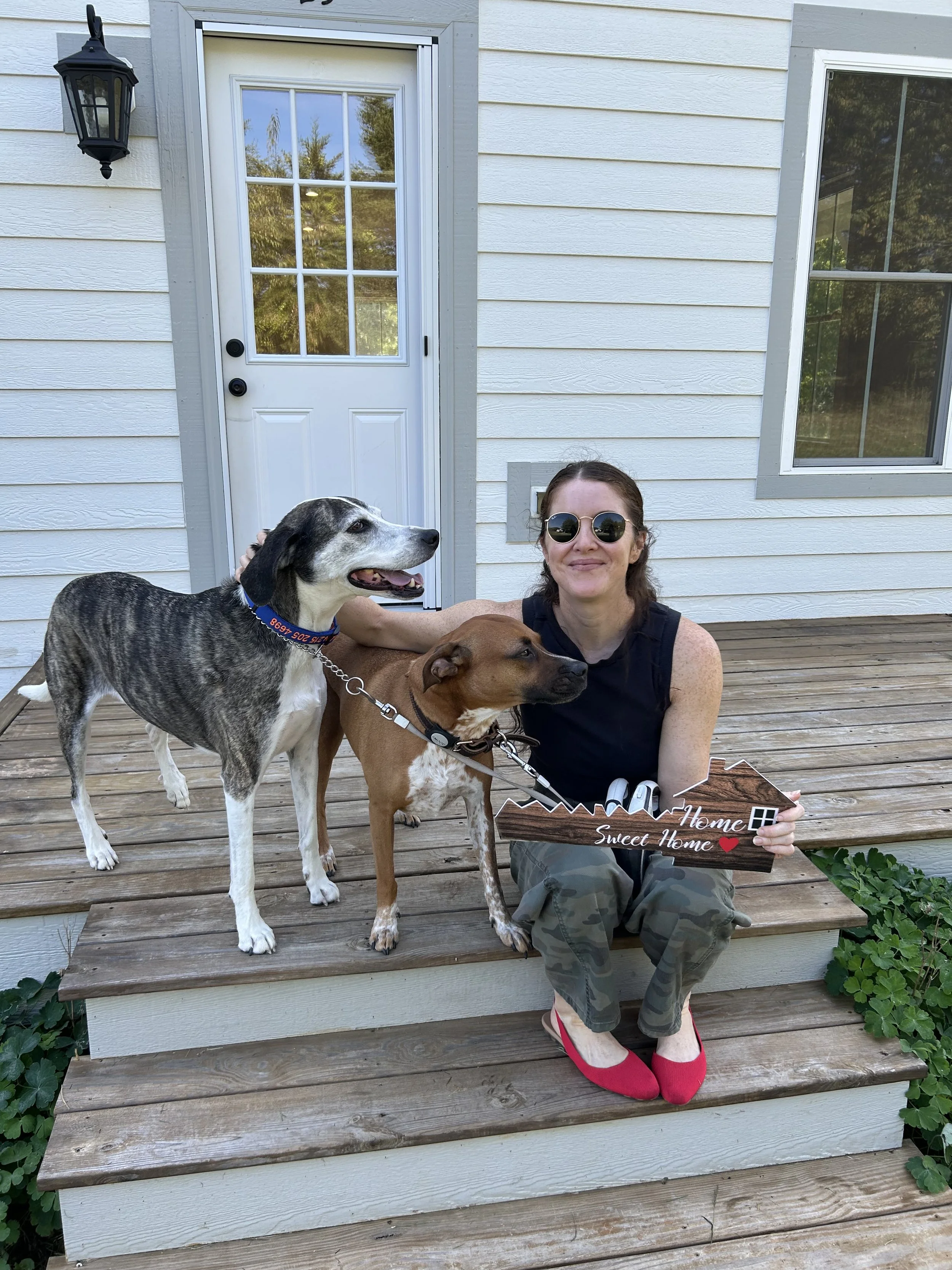 Client Julia, in sunglasses, hair pulled back, sitting on wooden stairs next to three happy dogs, holding a sign that says ‘Home Sweet Home’. The background shows a white house with a door and window, and a black outdoor lantern.