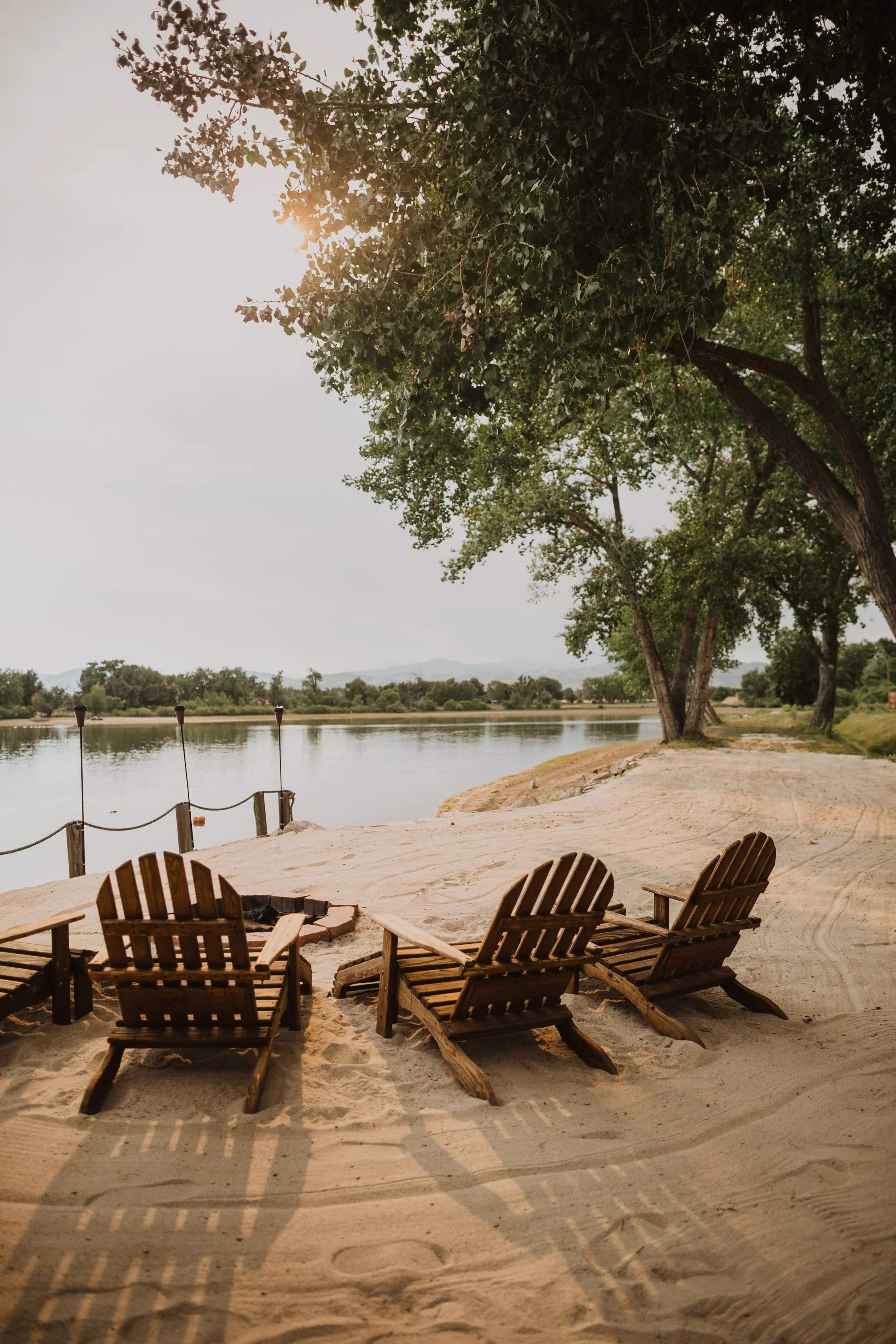 A lakeside scene with wooden chairs on sandy ground and tall trees providing shade. The sun is partially visible through the tree leaves, creating shadows on the sand. The lake waters are calm, and there are small buoys along the shoreline.
