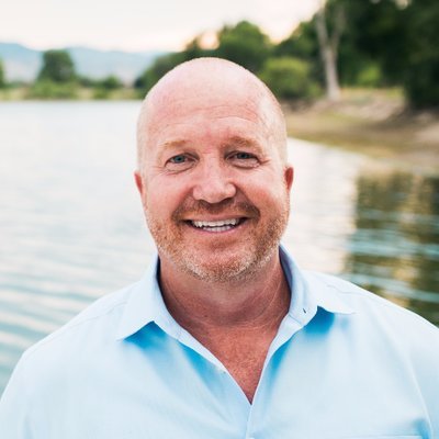 A smiling man with a bald head and beard, wearing a light blue shirt, standing outdoors near a body of water with trees and a grassy shoreline in the background.