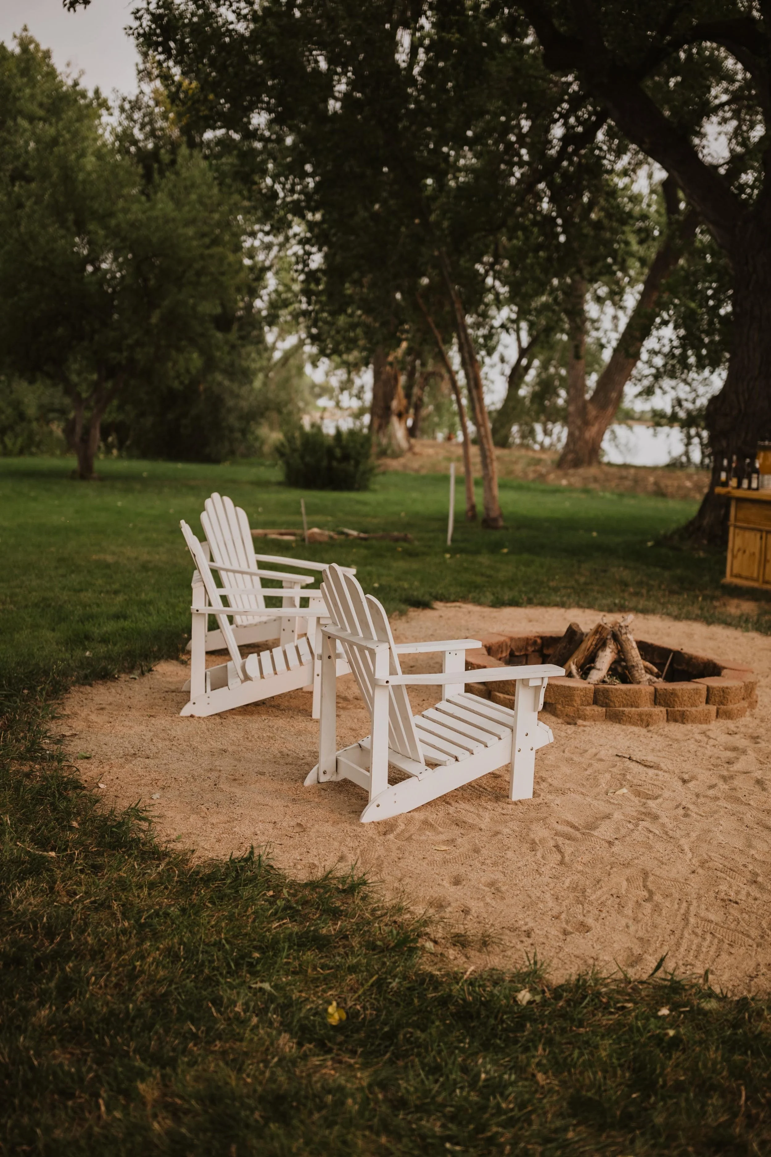 Two white Adirondack chairs facing a fire pit on sandy ground, surrounded by green grass and trees, with a lake or river in the background, during evening or late afternoon.