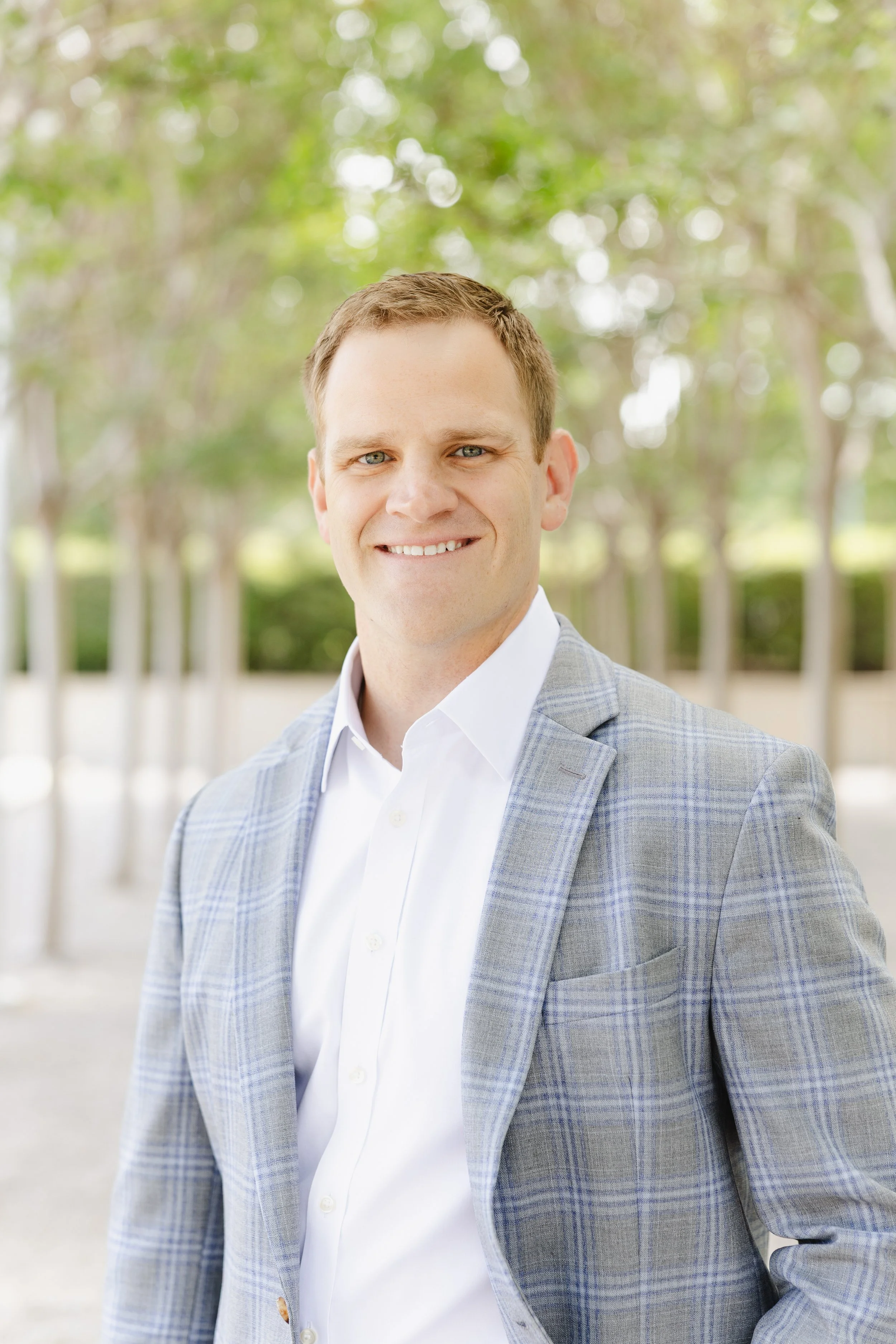 A smiling man wearing a light blue plaid blazer and white shirt, standing outdoors with a blurred green and white background of trees.