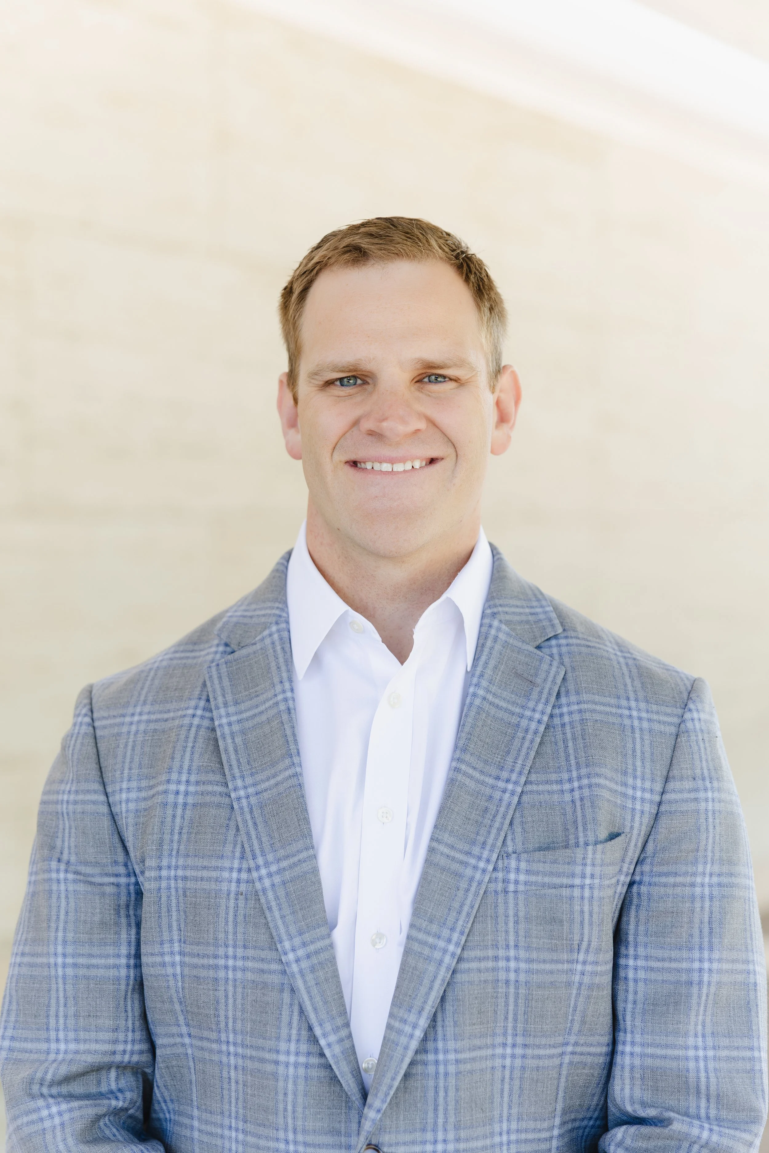 A man with short light brown hair and blue eyes smiling, wearing a light gray checkered blazer and a white dress shirt, against a light, neutral background.