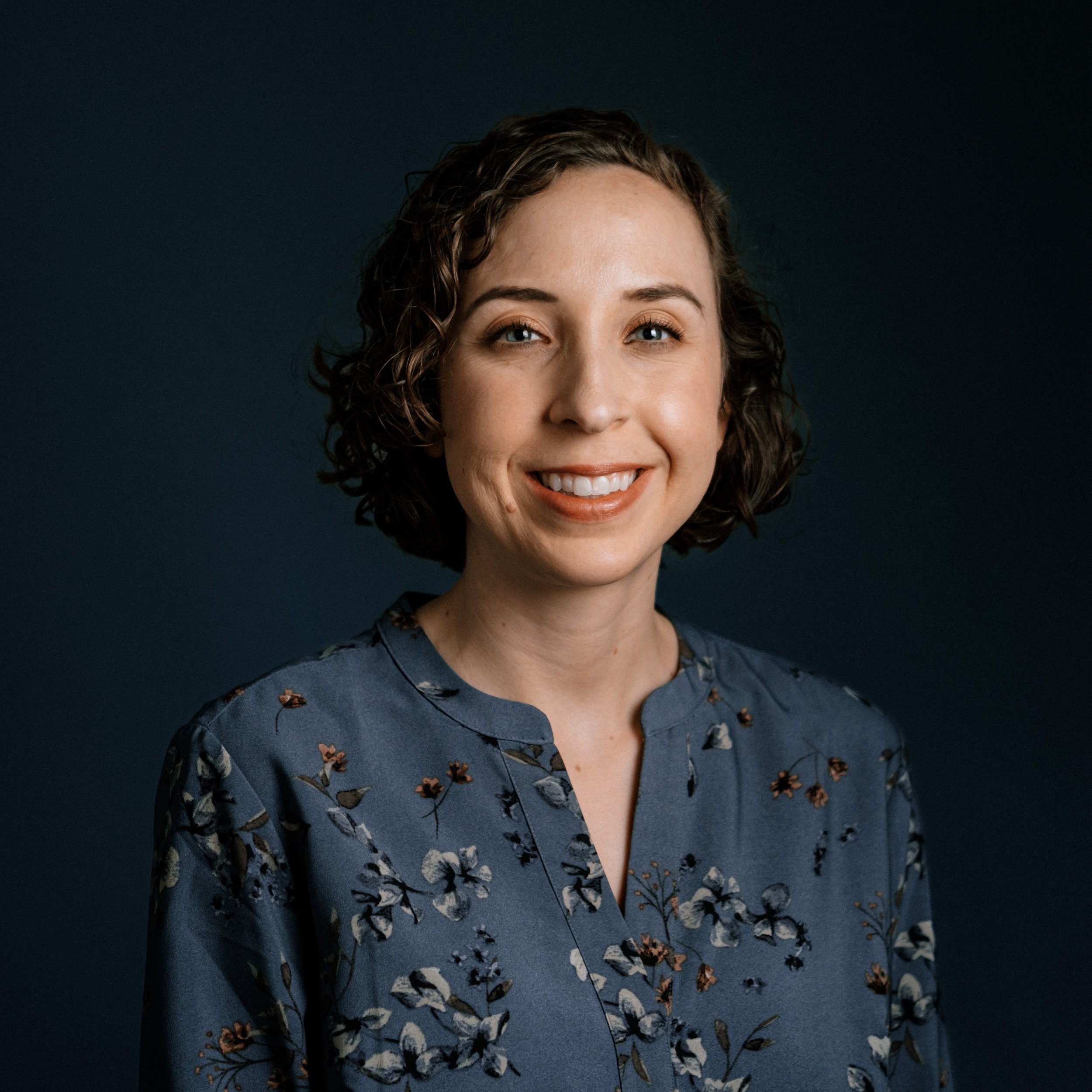 A woman with shoulder-length curly hair and light skin smiling, wearing a blue dress with a butterfly pattern, against a dark background.