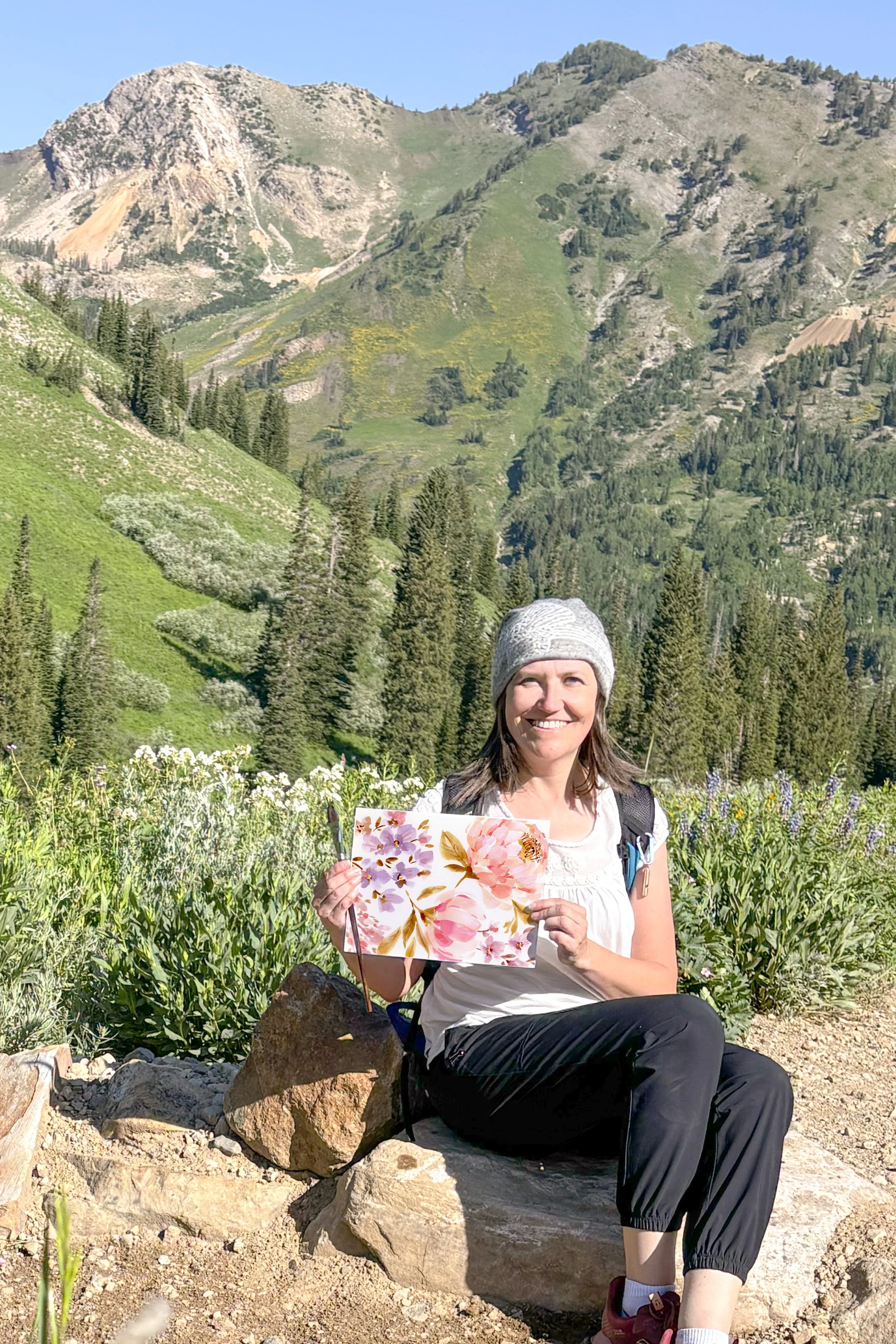 A smiling woman sitting on rocks in a field of tall green plants in front of mountain landscape, holding a floral-patterned paper.