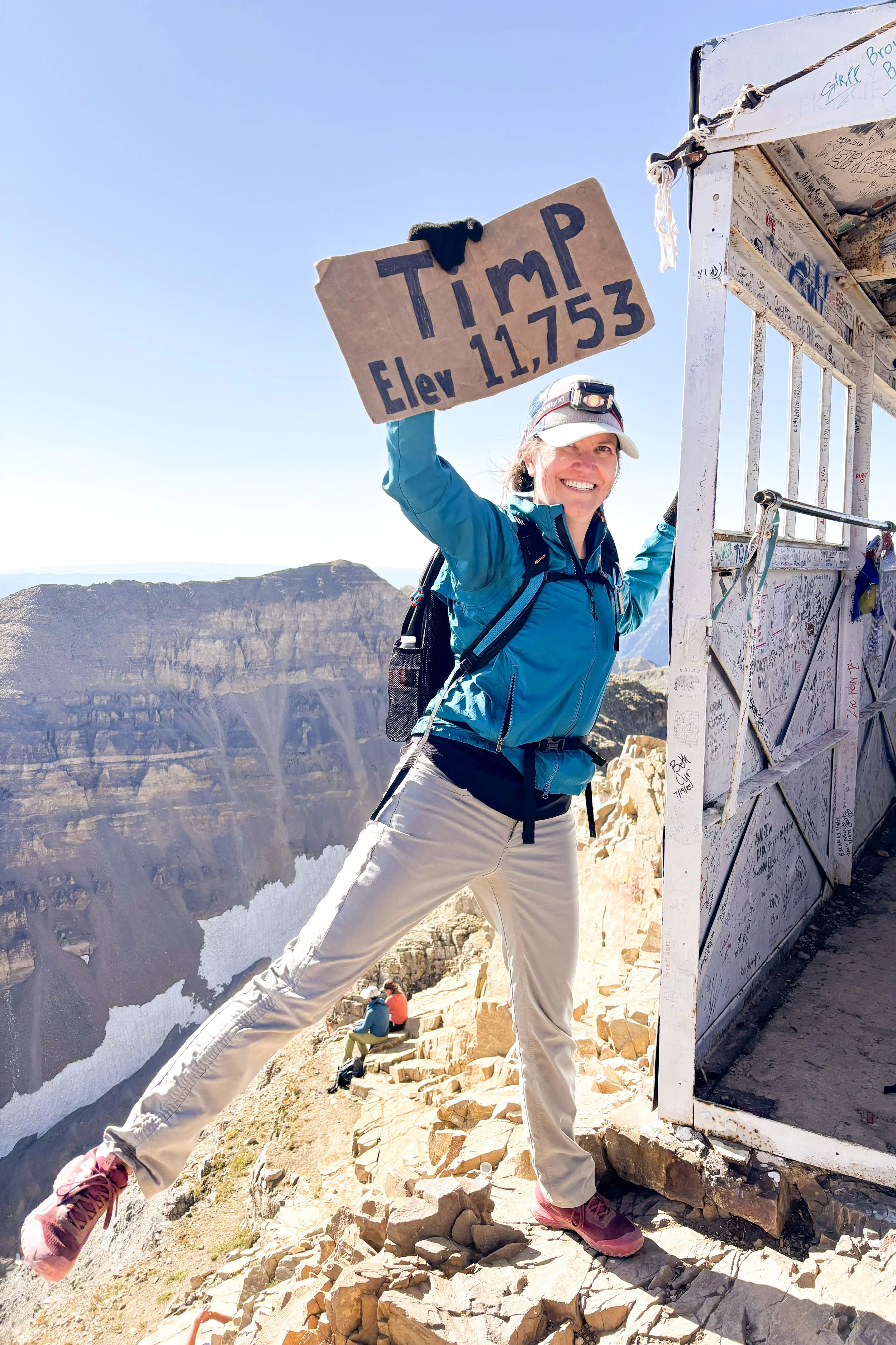A woman in outdoor hiking gear on a mountain trail holding a large cardboard sign with text, smiling at the camera, with a rocky landscape and a weather station or structure in the background.