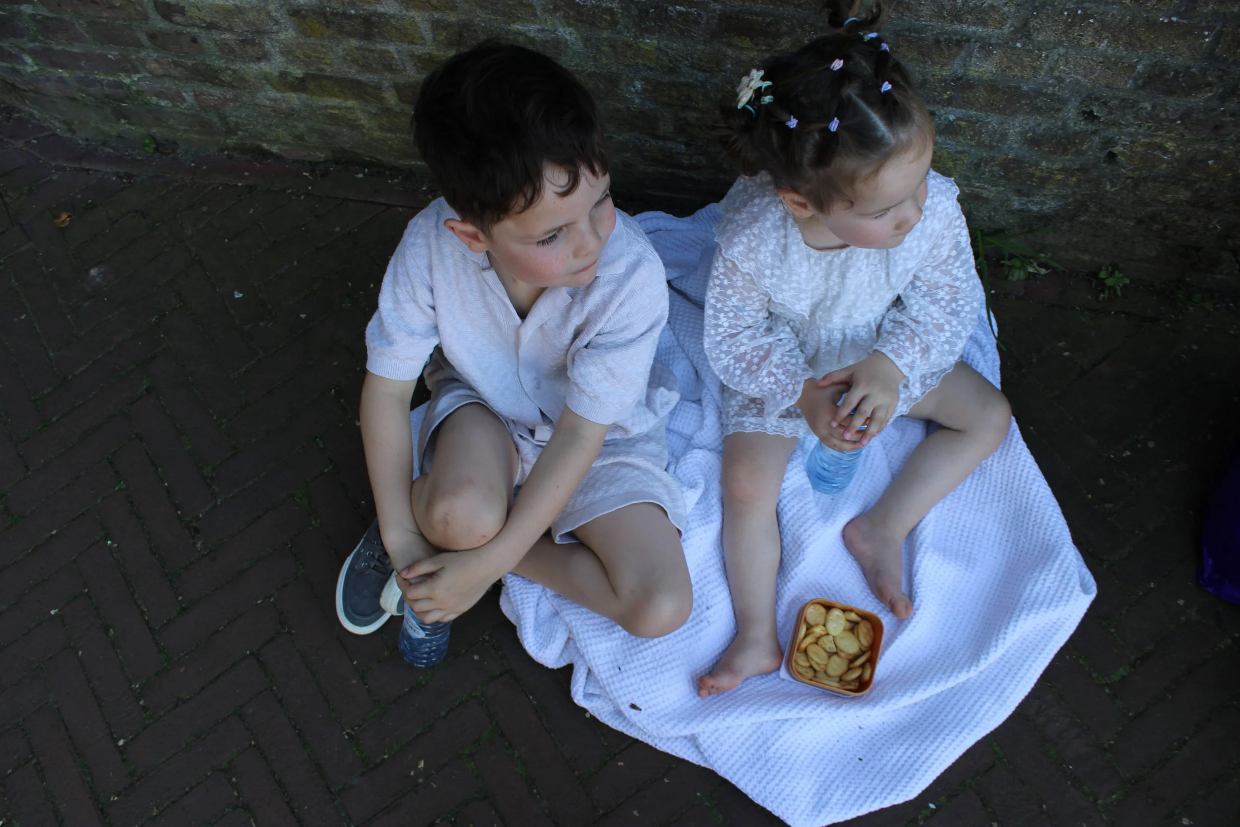 Two young children sitting outdoors on a blanket with a bowl of crackers, near a brick wall.