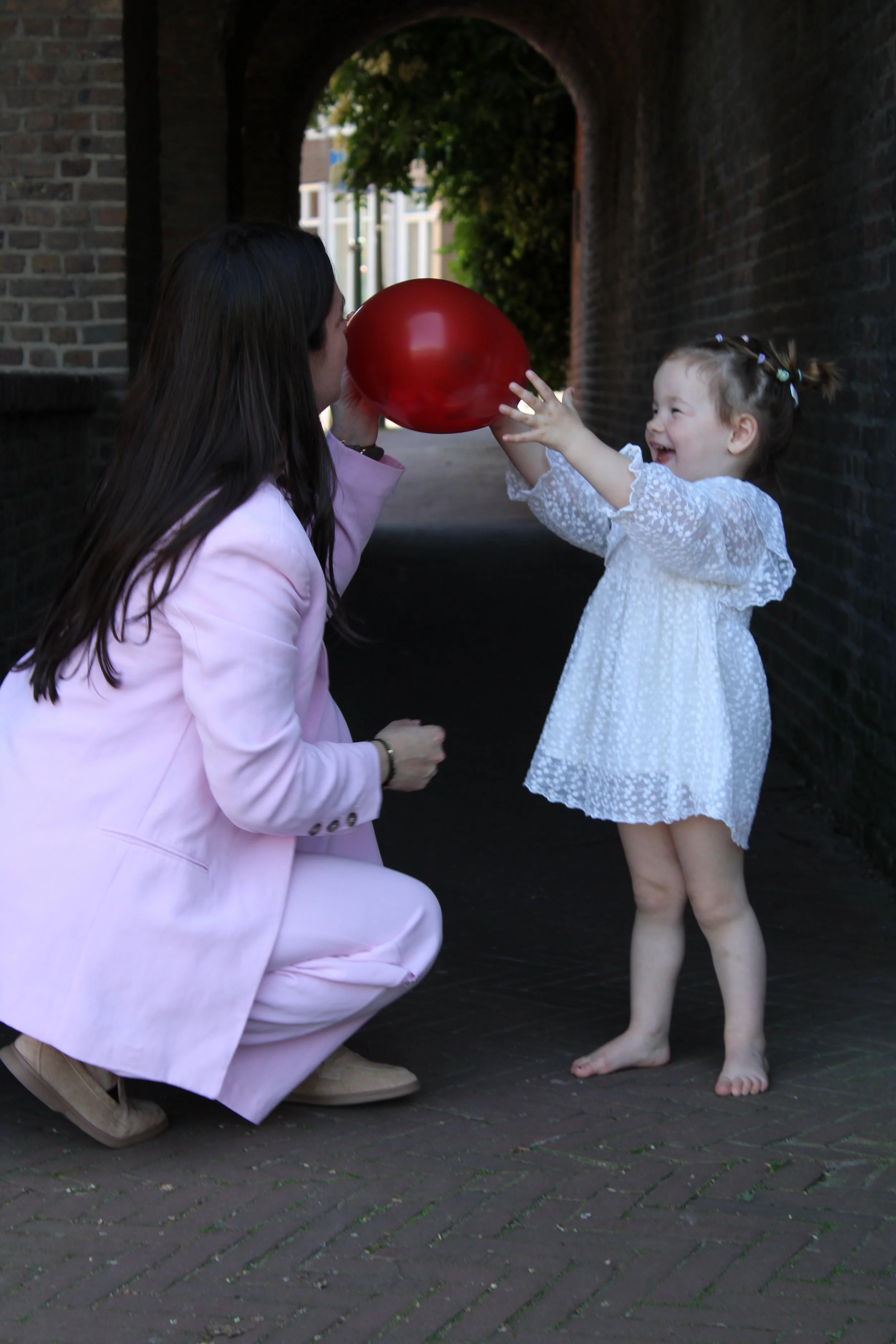 A woman in a pink suit kneeling and holding a red balloon in front of a young girl with curly hair in a white dress, under a brick archway.