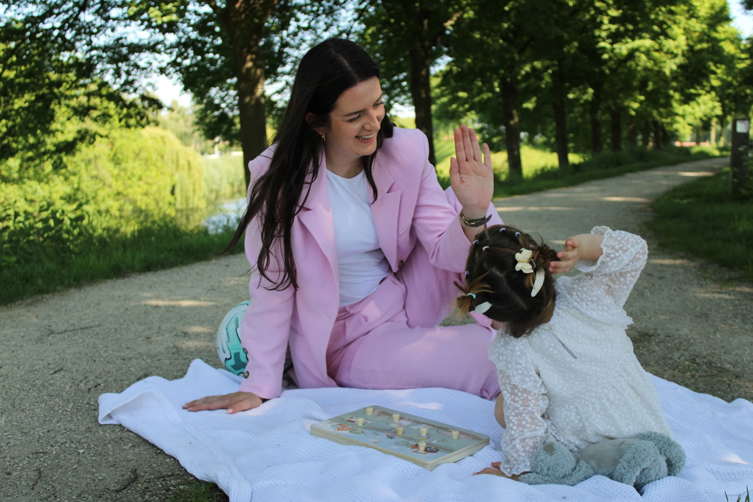 A woman in a pink blazer and a girl in a white lace dress are playing checkers on a white blanket outdoors near trees.