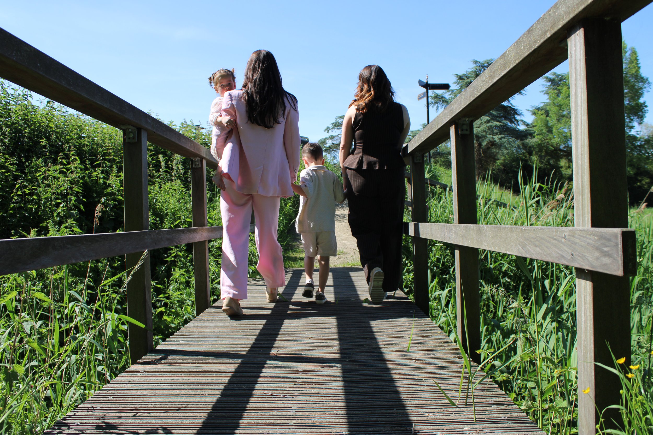 Four people, including two children and two women, walking on a wooden footbridge over a green area on a sunny day.