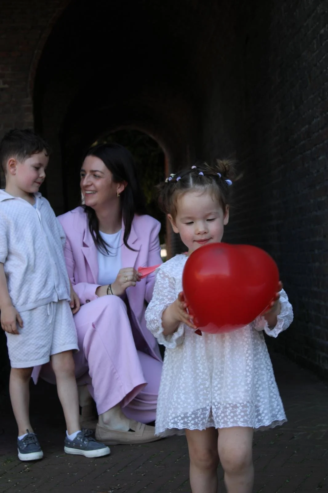 A woman with two young children, one holding a heart-shaped red balloon, in a dark tunnel.