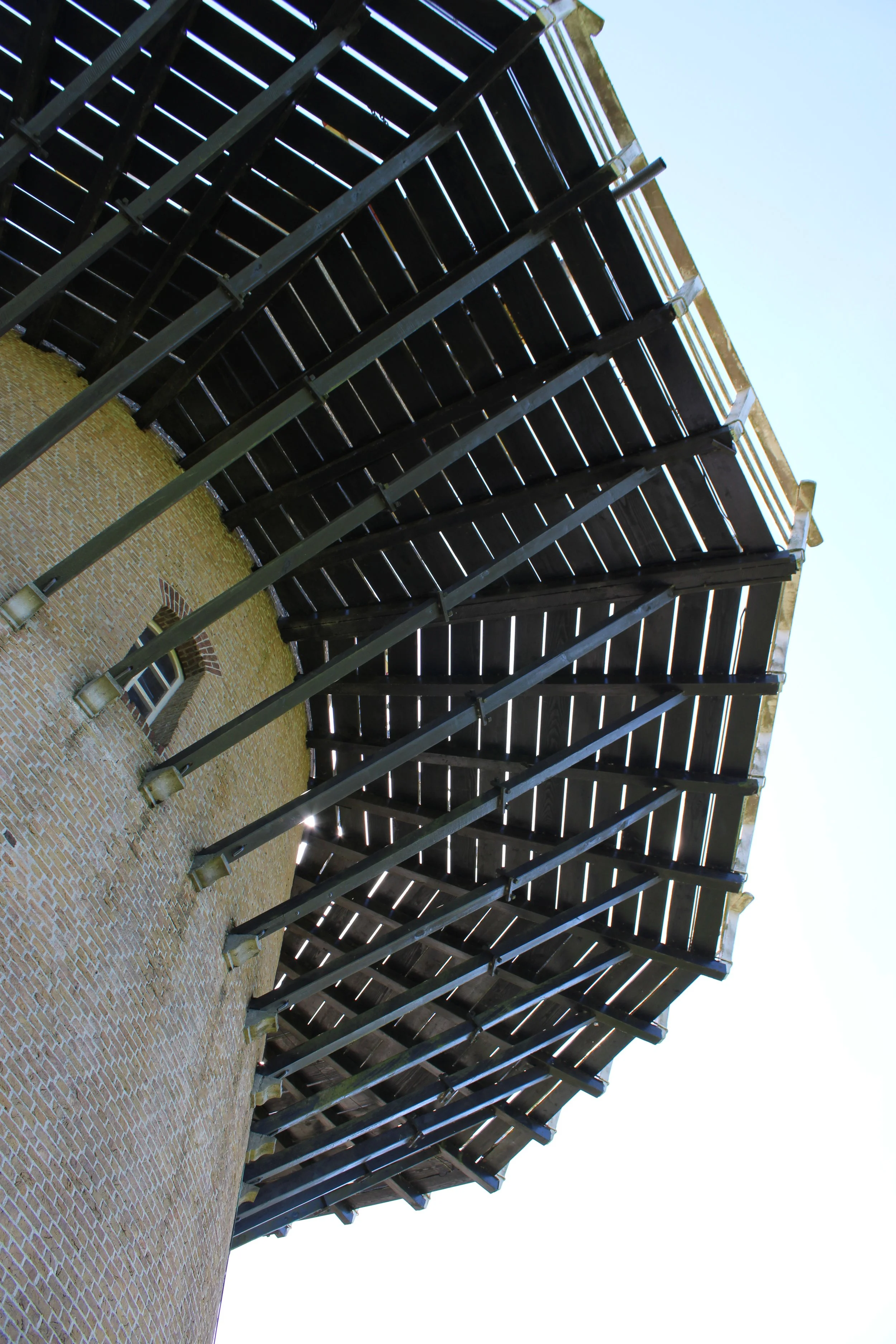 The image shows the underside of a multi-level wooden outdoor staircase with metal supports against a brick building wall, with a clear blue sky in the background.
